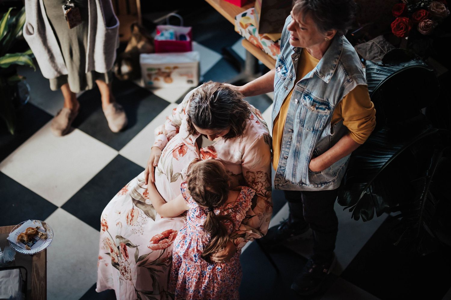 Guests hug and embrace a pregnant woman at a baby shower in a cozy shop with checkered floors.