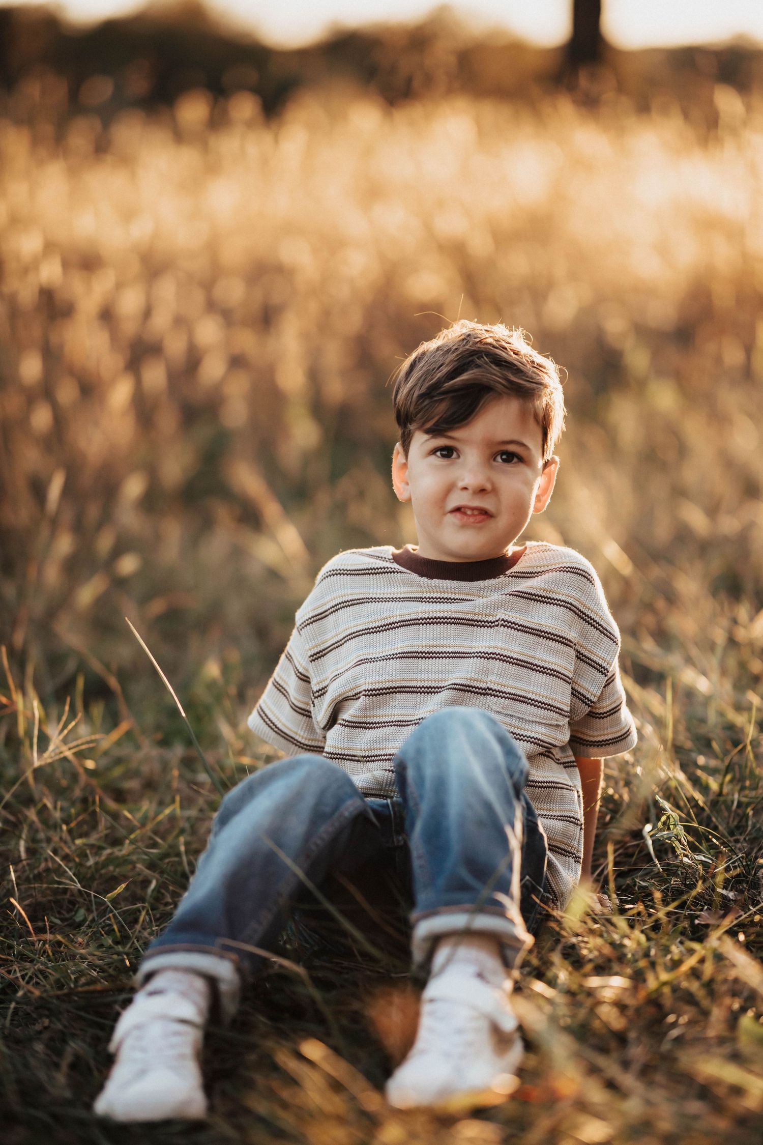 A young boy in a striped shirt and jeans sits in a golden sunlit field, smiling at the camera.