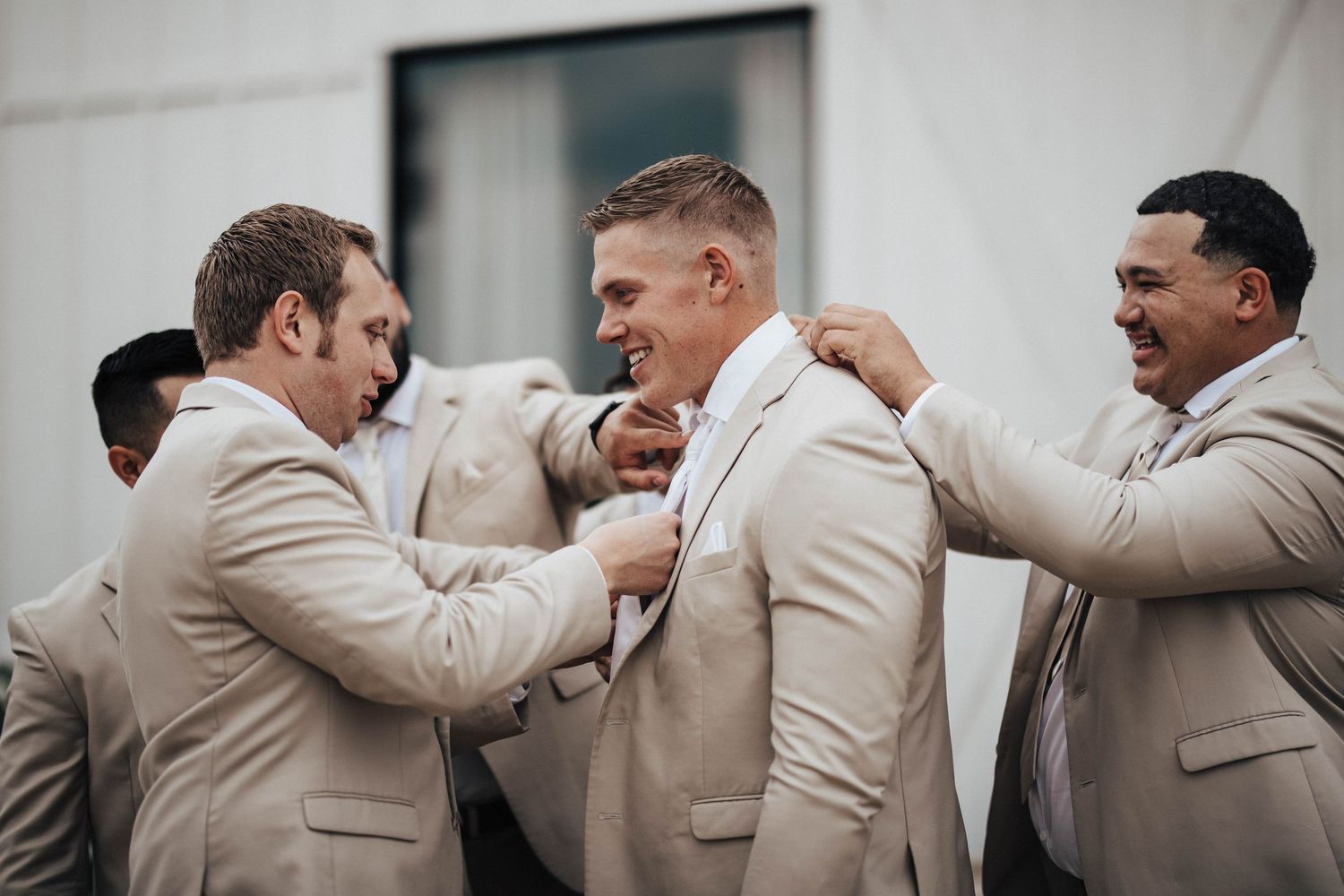 Groomsmen in tan suits laughing and adjusting the grooms tie before the wedding ceremony.