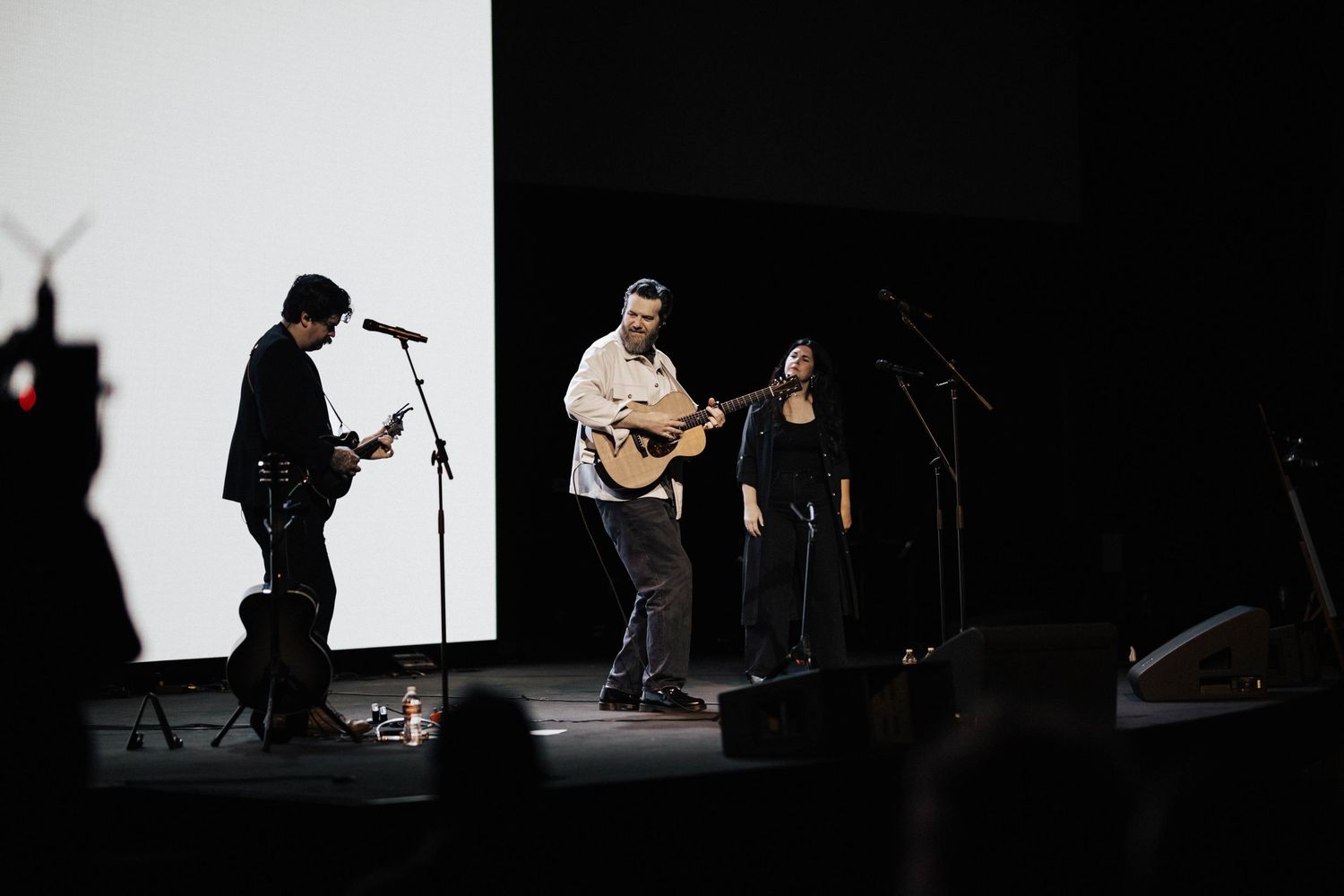Three musicians perform together on stage with acoustic guitar in front of large white projection screen.