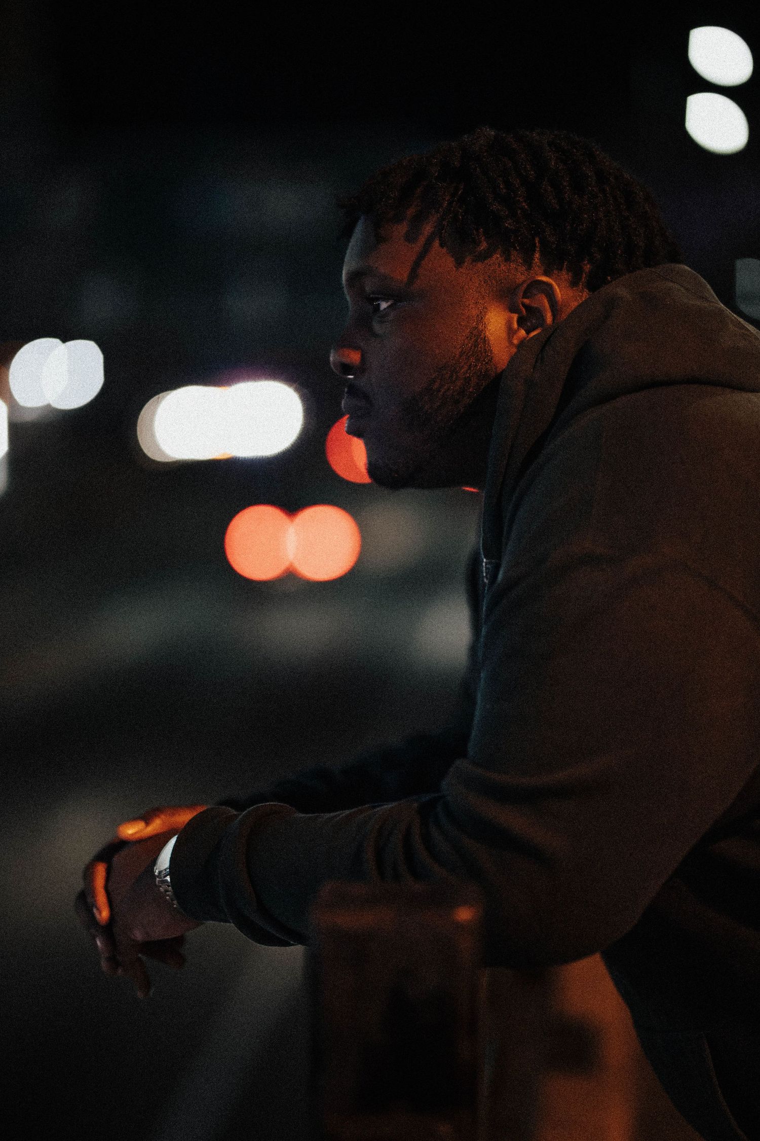 Young Black man in a hoodie leaning on a railing at night with bokeh city lights behind him.