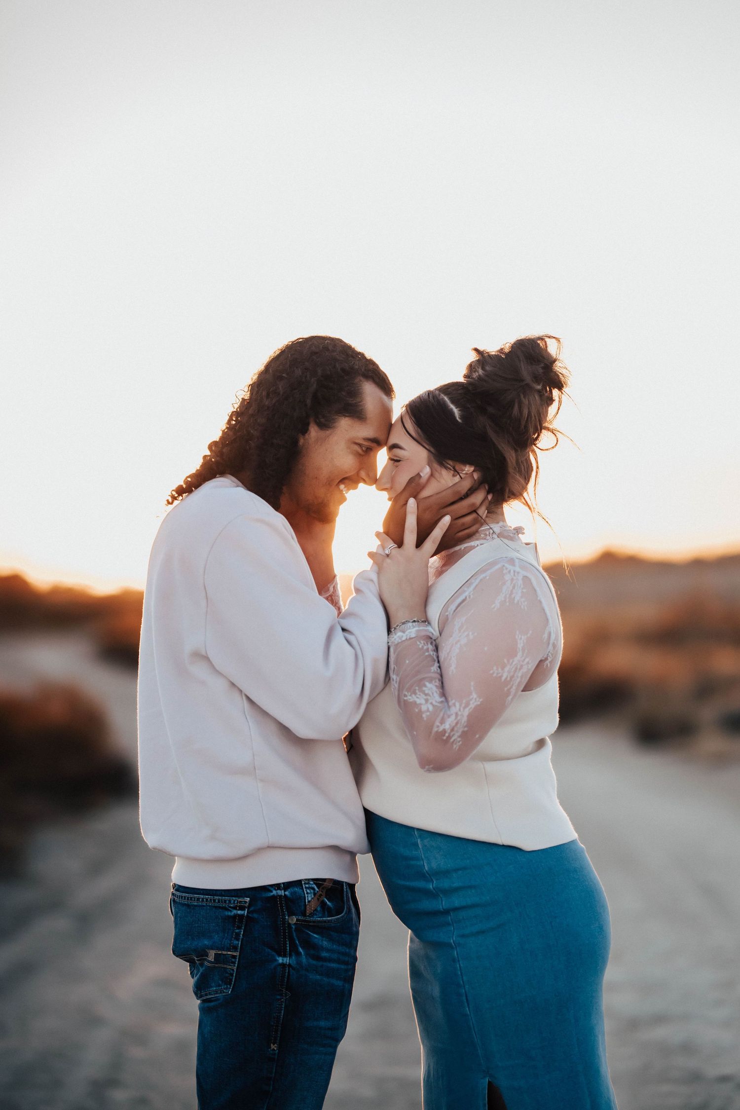 Romantic couple touching foreheads tenderly at sunset on an open road, both wearing white tops and jeans.