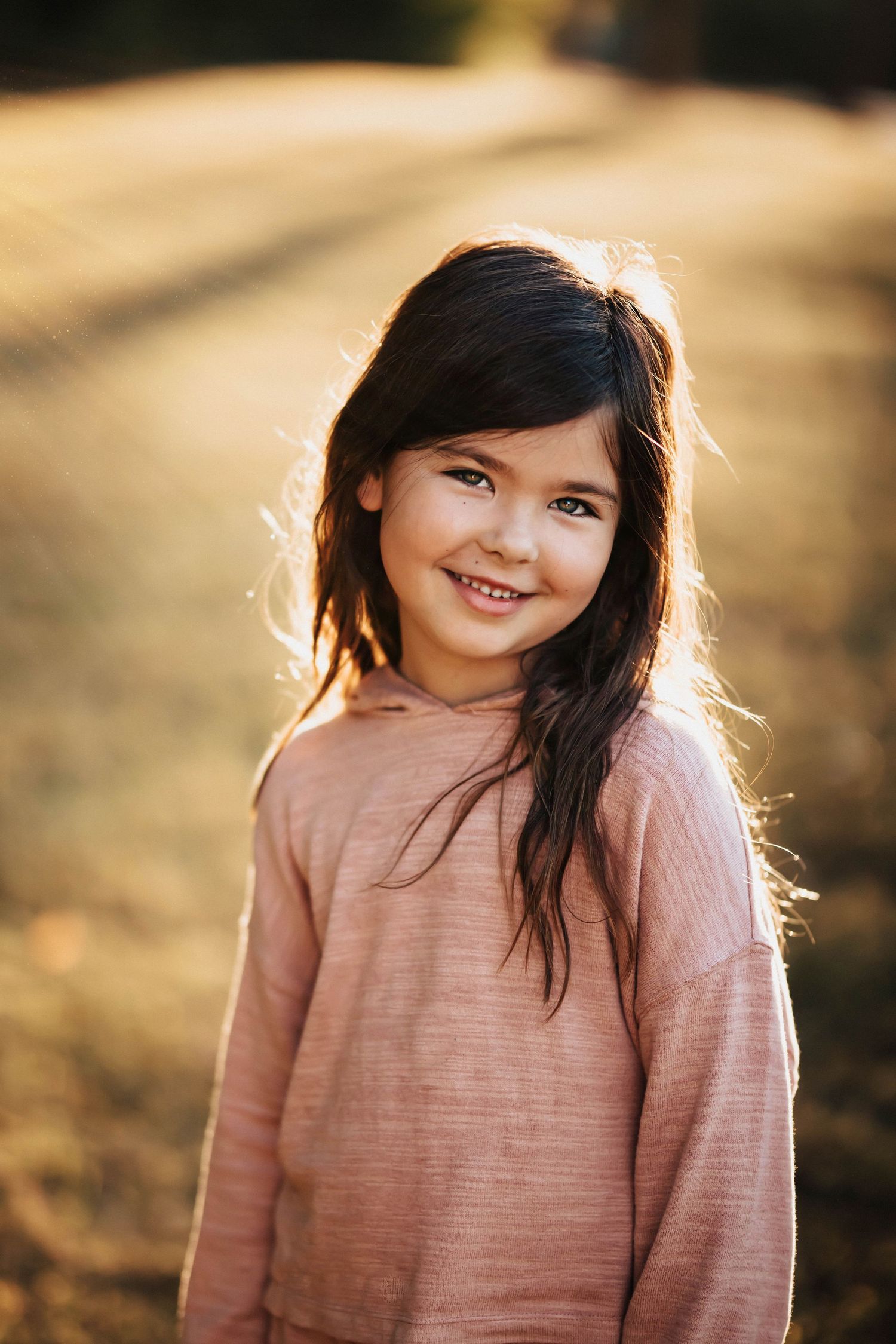 Smiling young girl with dark hair in pink hoodie standing in sunlit field during golden hour.