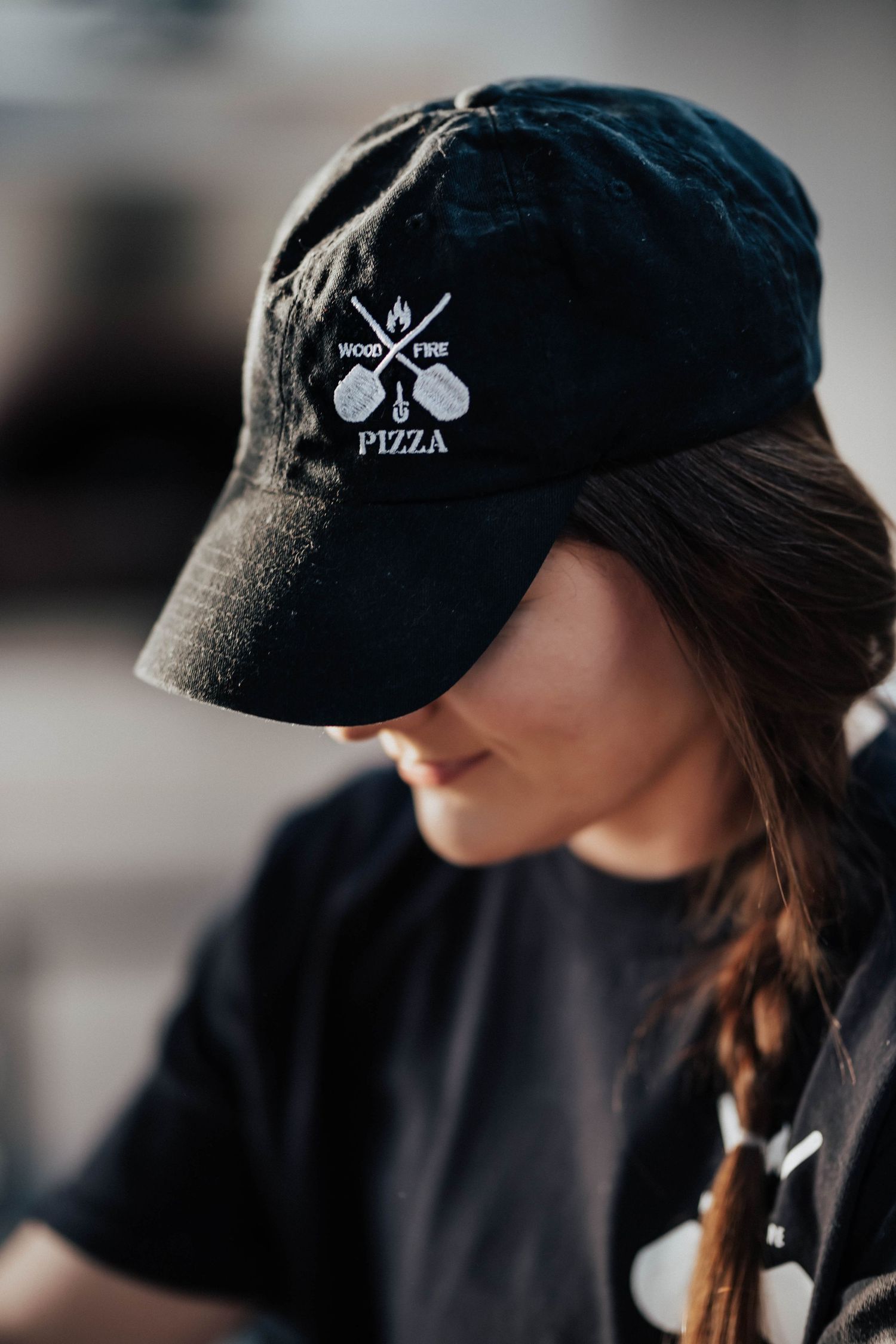 Woman wearing a black Wood Fire Pizza embroidered cap with crossed pizza peels logo.