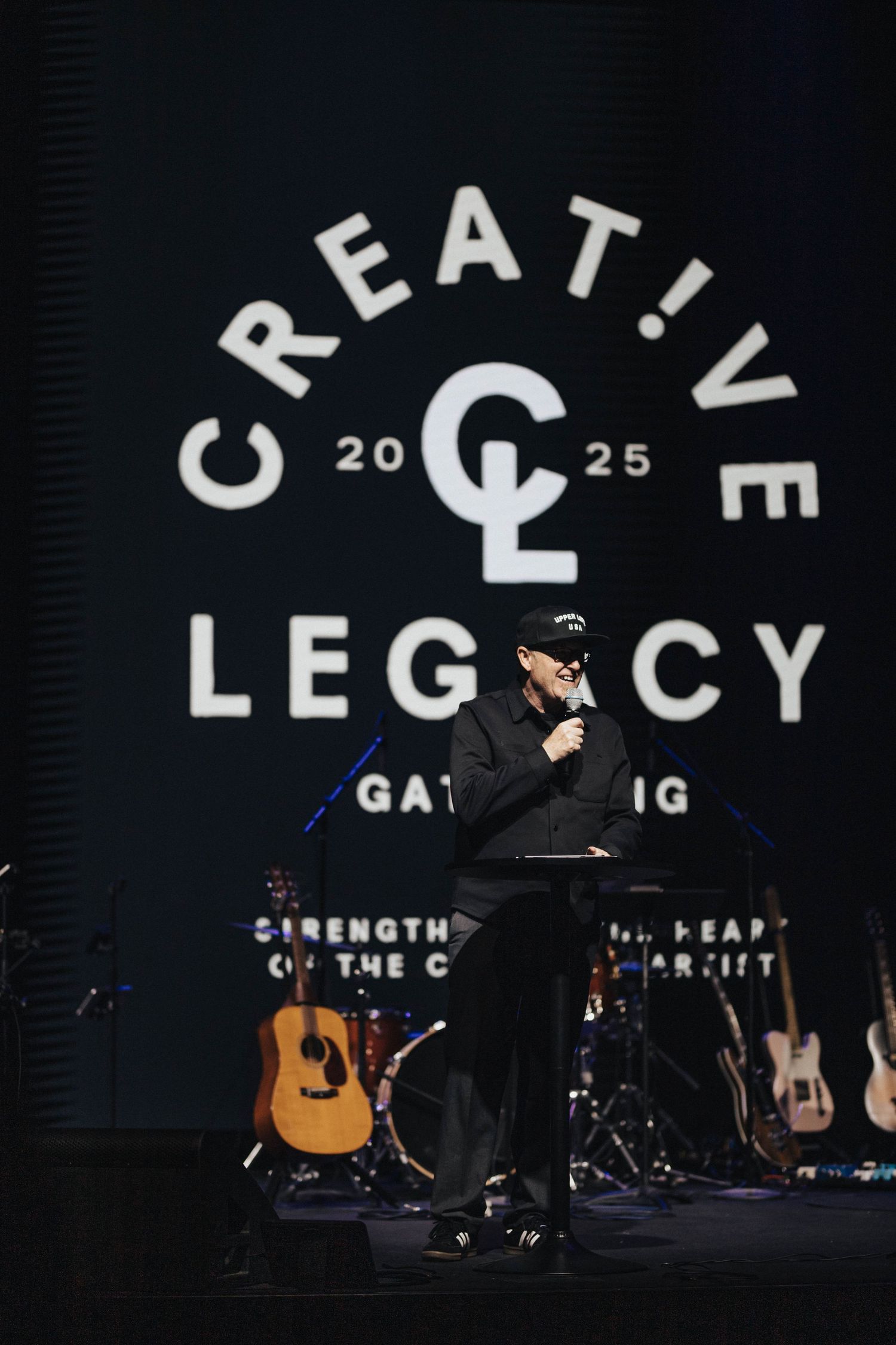 Speaker at podium on stage at Creative Legacy Gathering 2025 event with logo projected on screen behind.