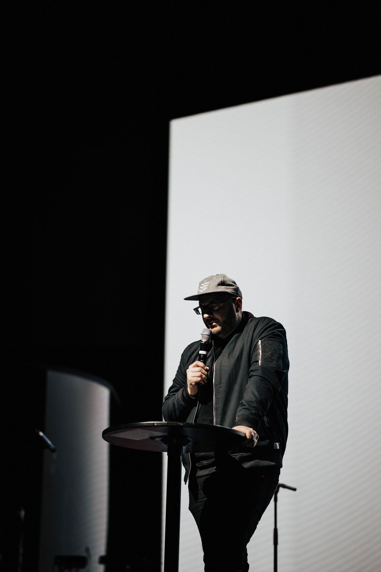 Man in cap and black jacket speaks into microphone at podium on dark stage with white screen.