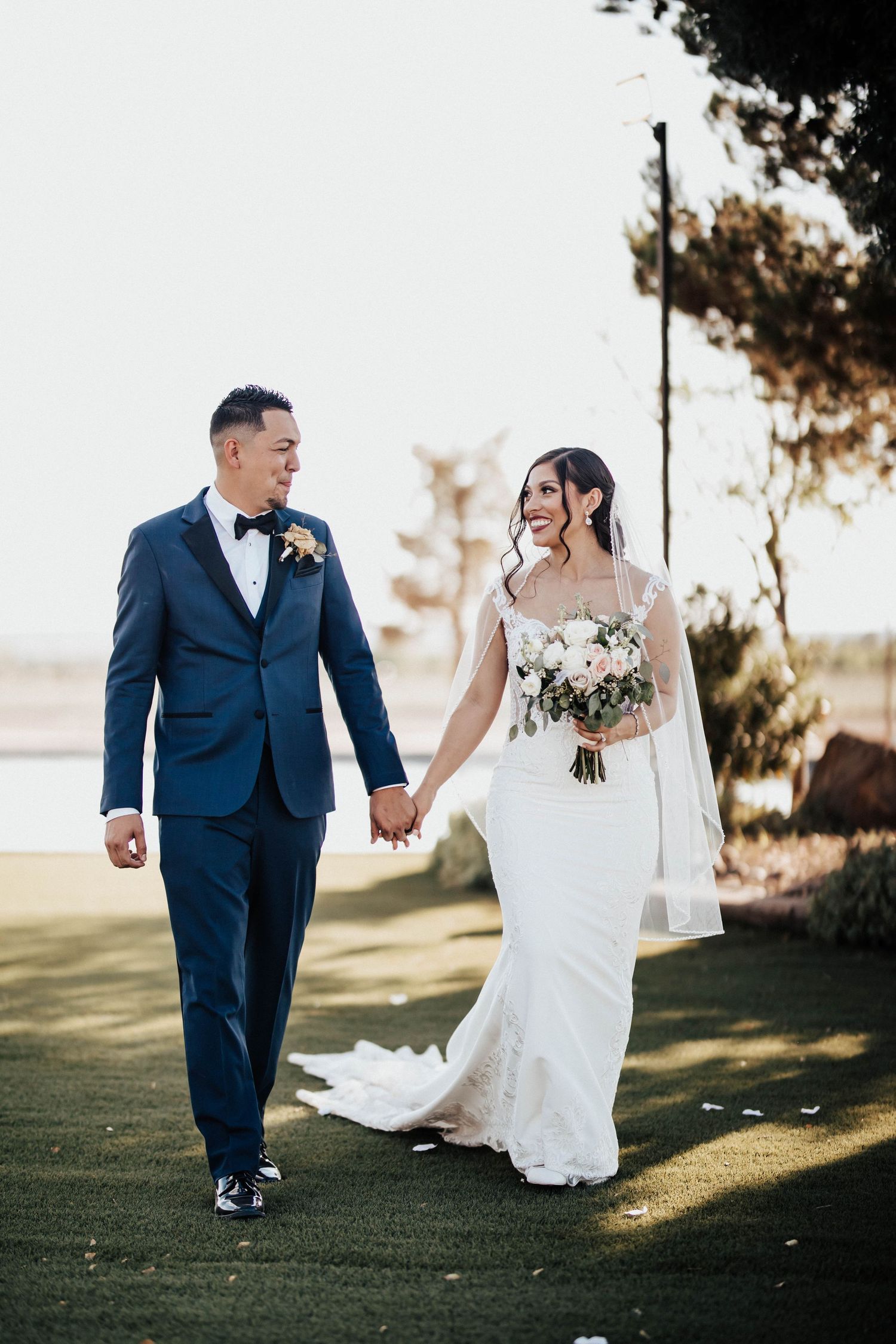 A smiling bride in a white gown holding a bouquet walks hand-in-hand with a groom in a navy suit on a green lawn.