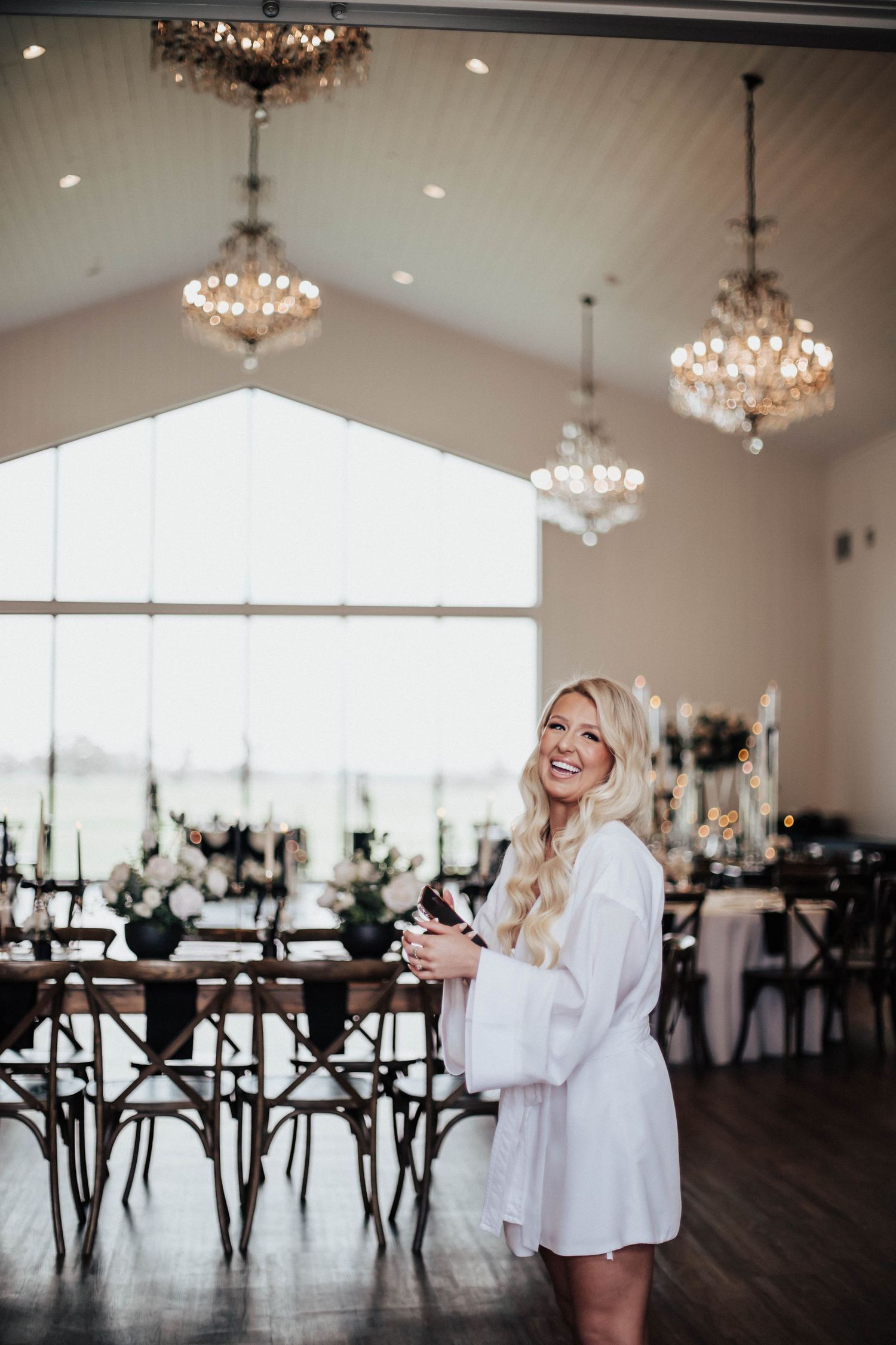 Smiling blonde bride in white robe holds bouquet in elegant chandelier-lit wedding venue with dark wood chairs.