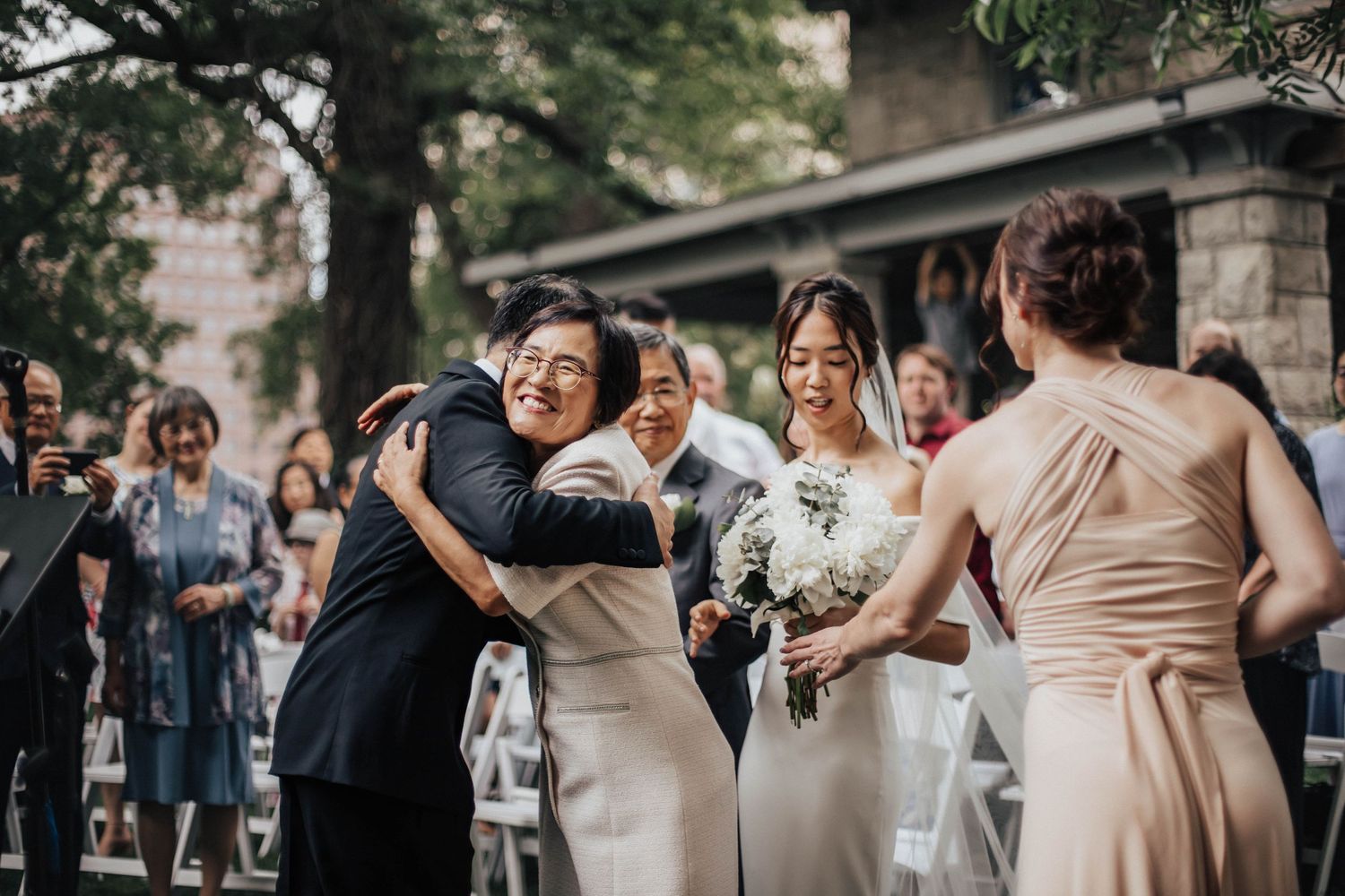 Bride in white dress hugs guest during outdoor wedding ceremony while bridesmaid in champagne gown stands nearby.