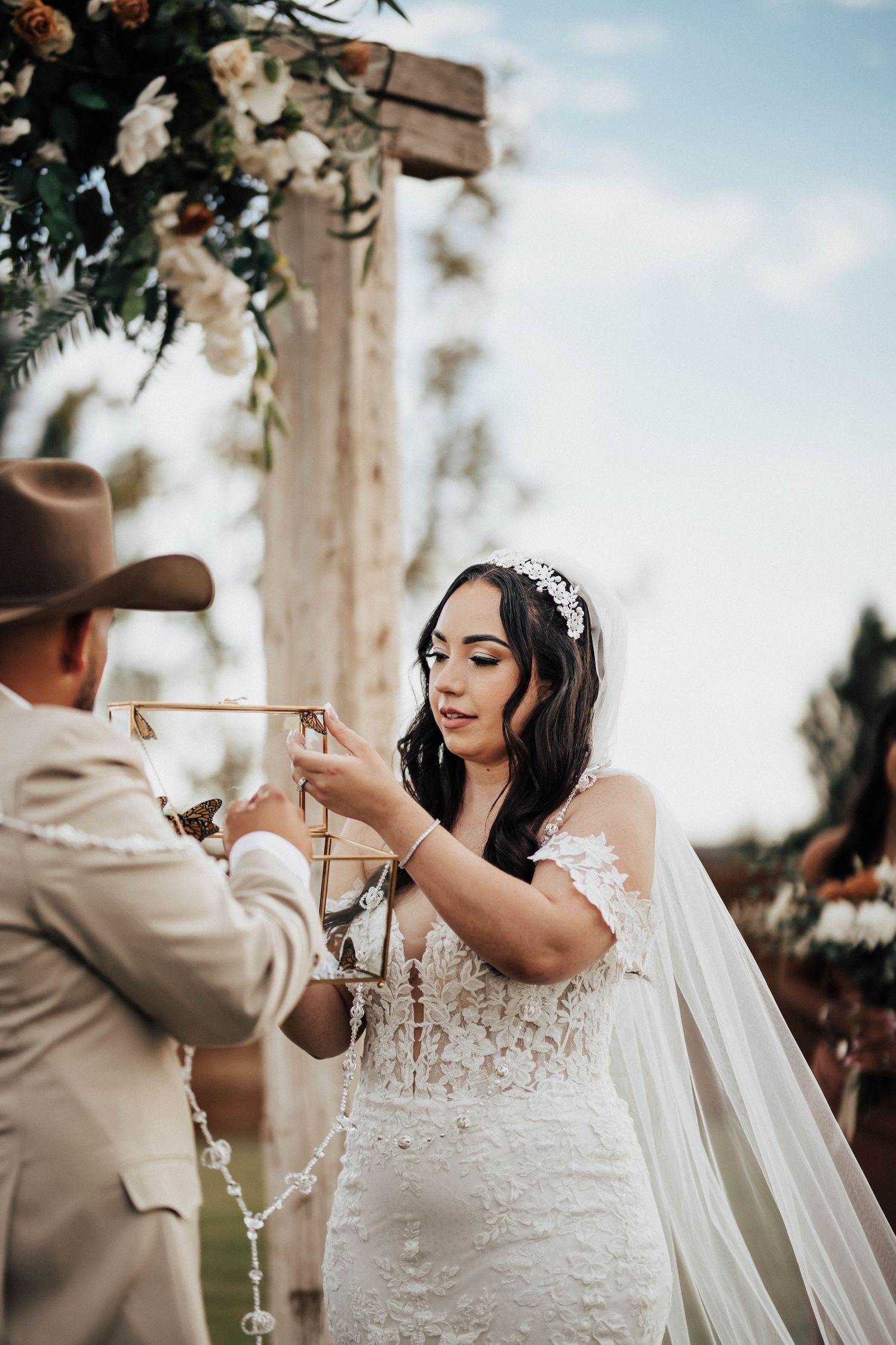 Bride in lace off-shoulder gown and veil receives wedding ring from groom in tan cowboy hat at outdoor ceremony.