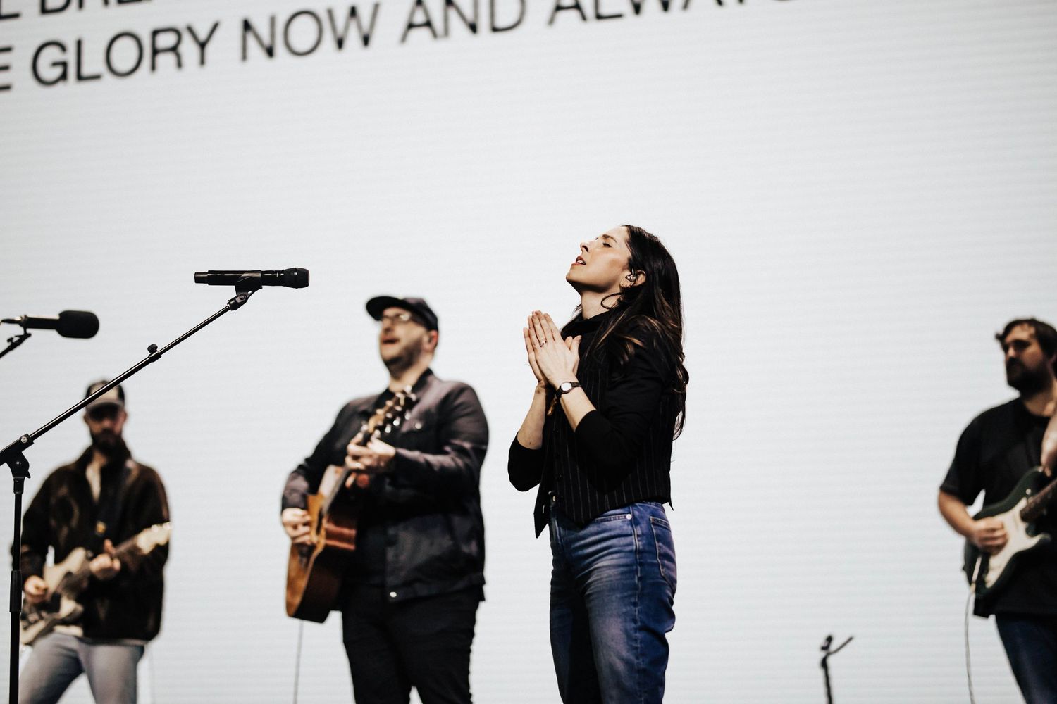 Female worship leader praying on stage with guitarists performing behind her during live service.