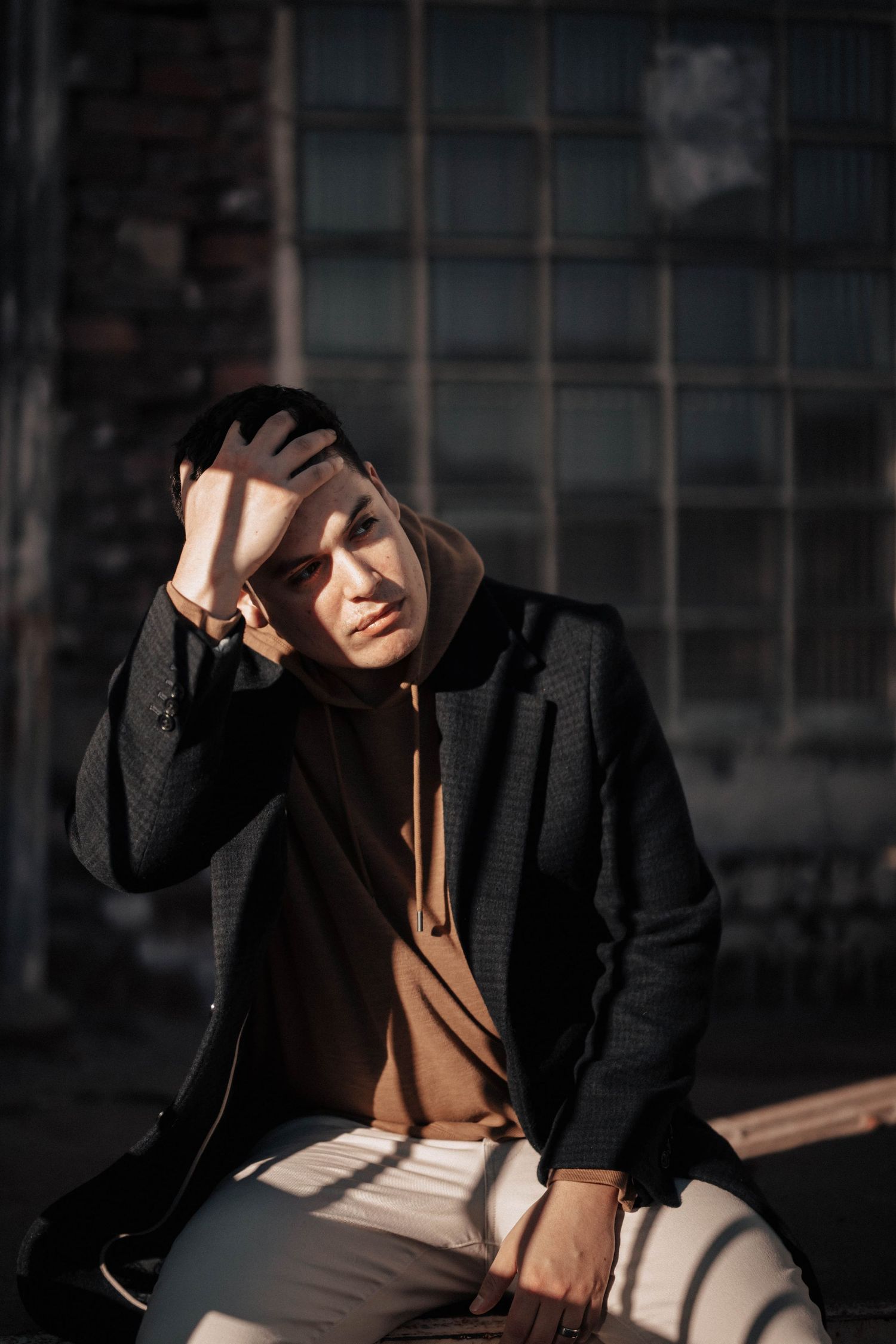 Young man in a black blazer and brown hoodie sitting outdoors shielding his eyes from sunlight.