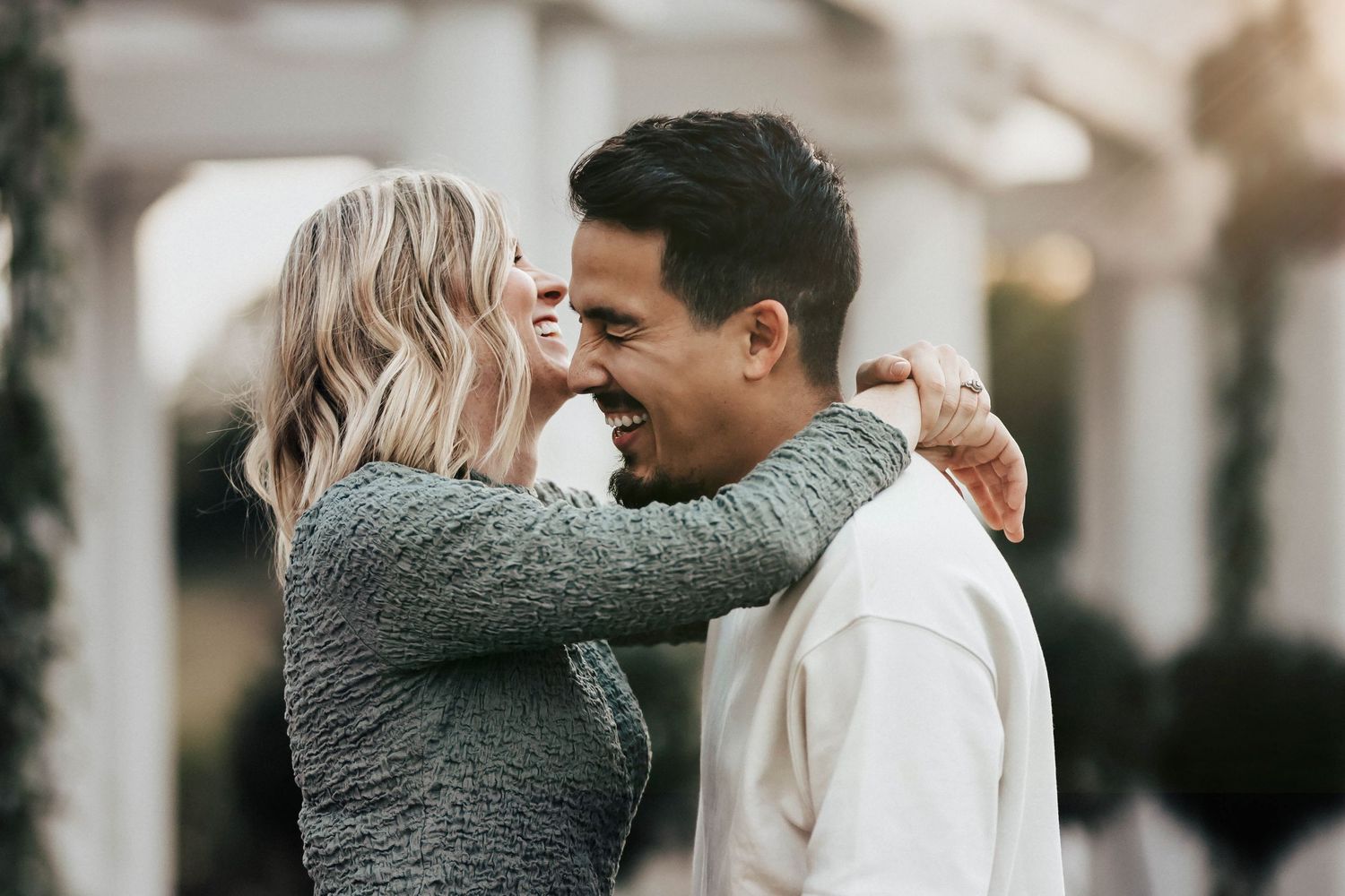 Joyful couple laughing and embracing outdoors, blonde woman in gray sweater hugging smiling man.