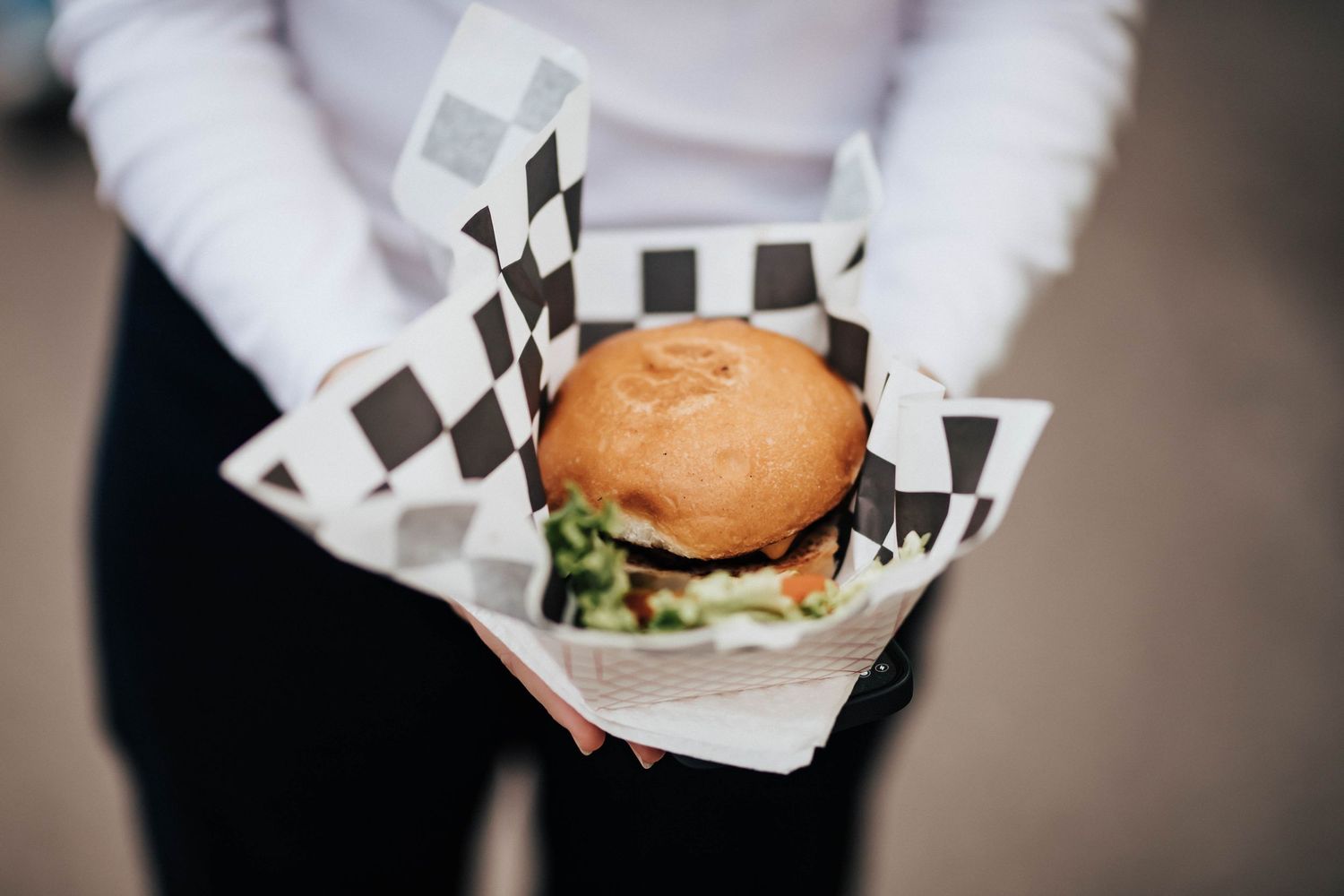 A person holds a burger in checkered paper tray at an outdoor event.