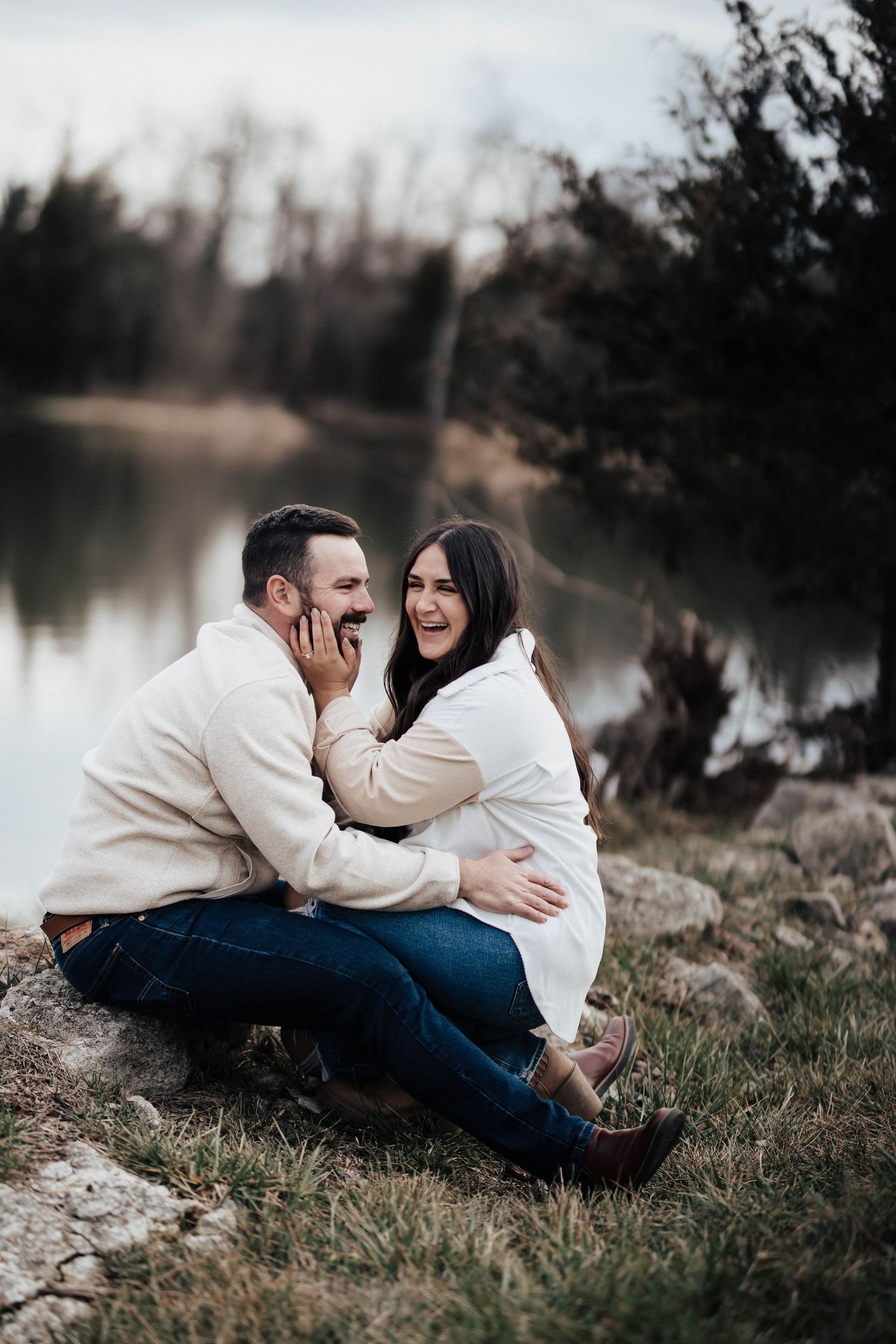 Couple sitting by a lakeside, laughing and embracing, wearing white sweaters and jeans in a serene outdoor setting.