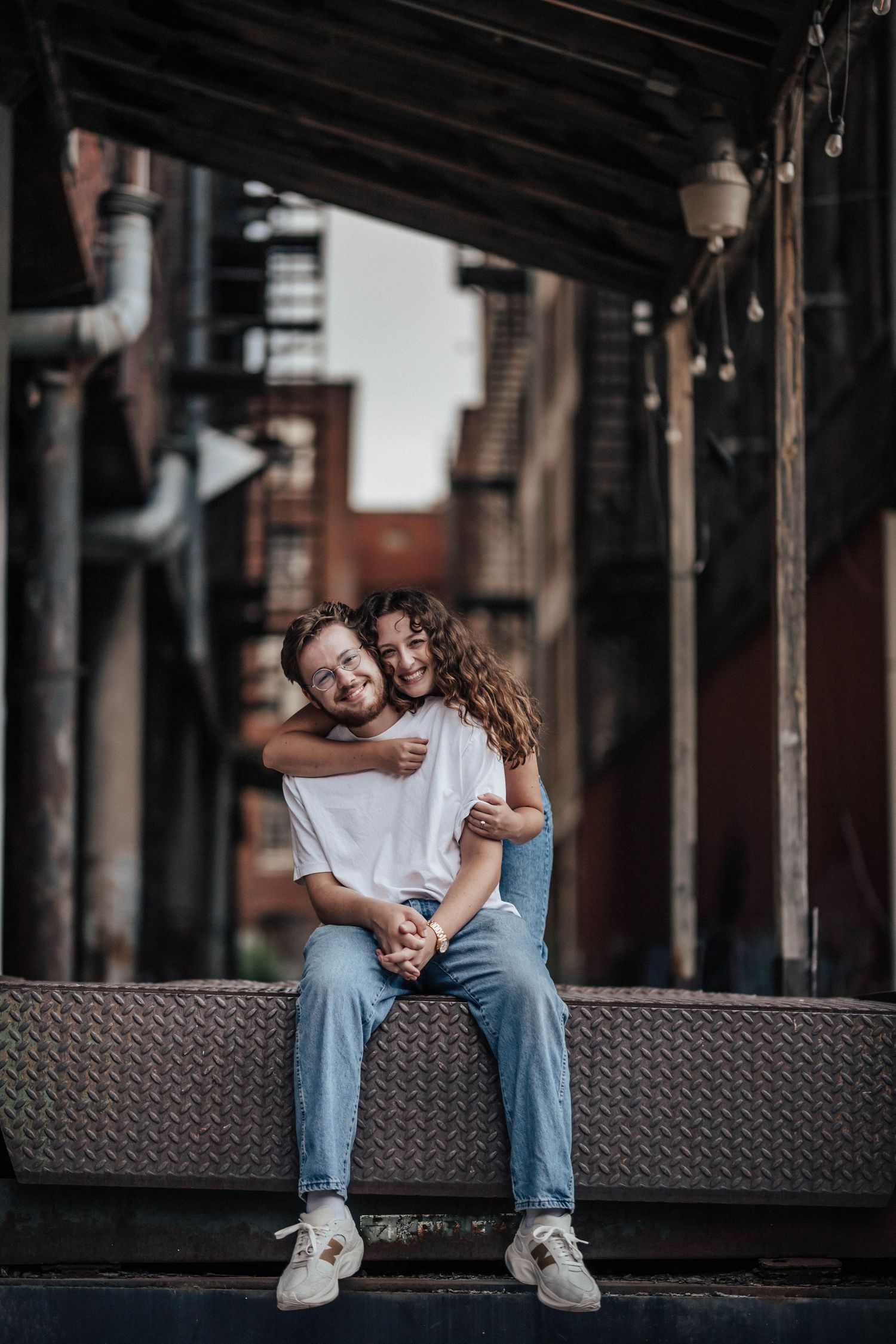 A couple sitting on a metal ledge in an urban alley, woman hugging man from behind, both wearing jeans and white sneakers.