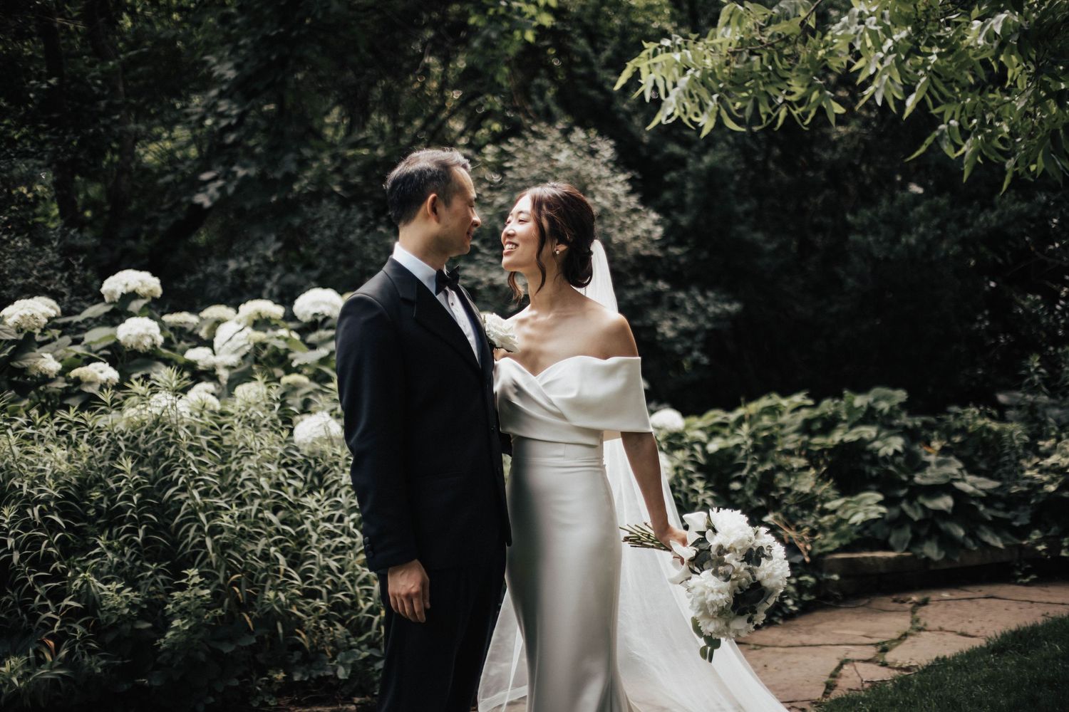 Bride and groom share a romantic moment in a lush garden, bride in off-shoulder gown holding white bouquet.