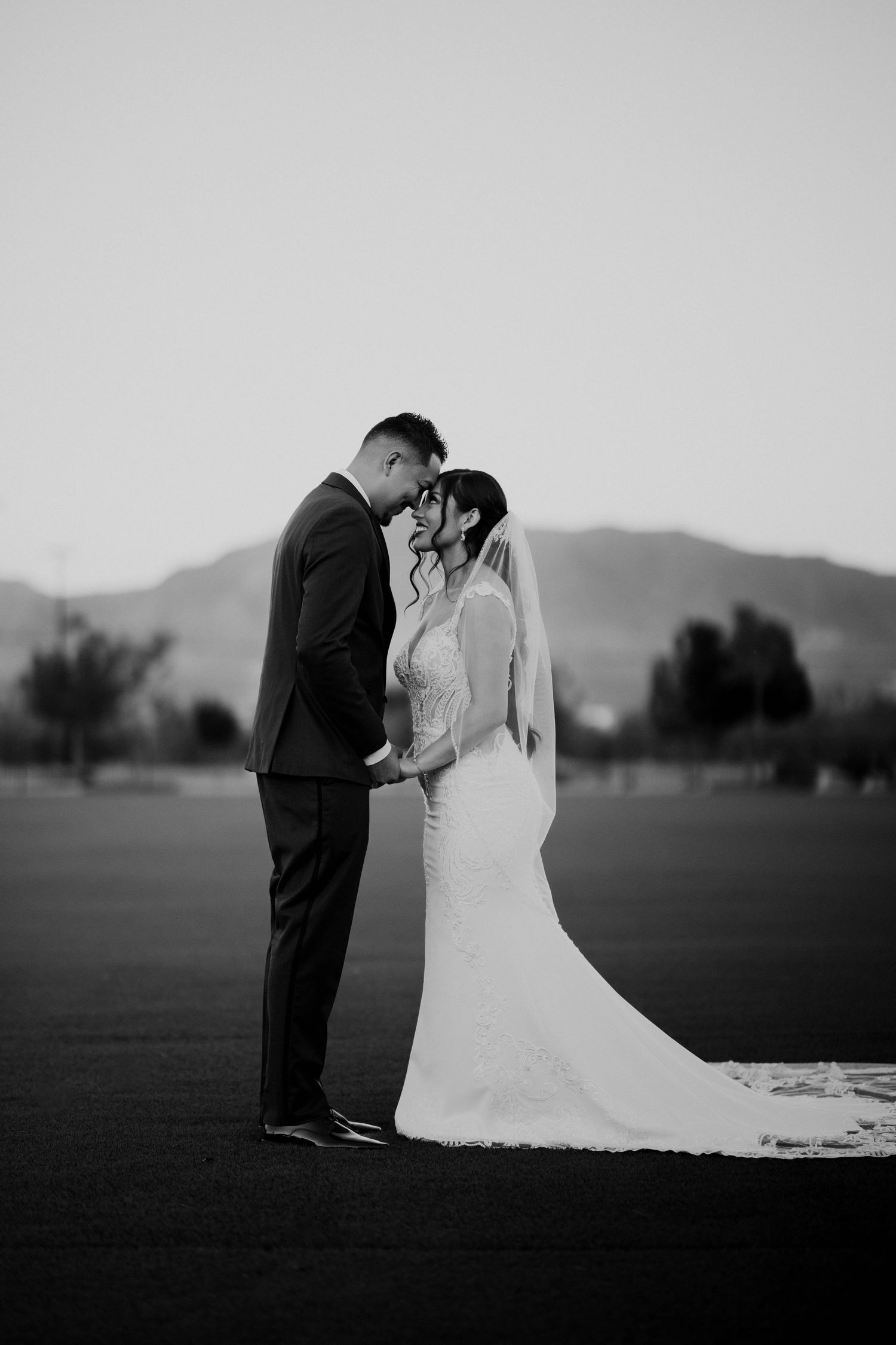 Black and white romantic portrait of bride and groom touching foreheads outdoors with mountain backdrop.