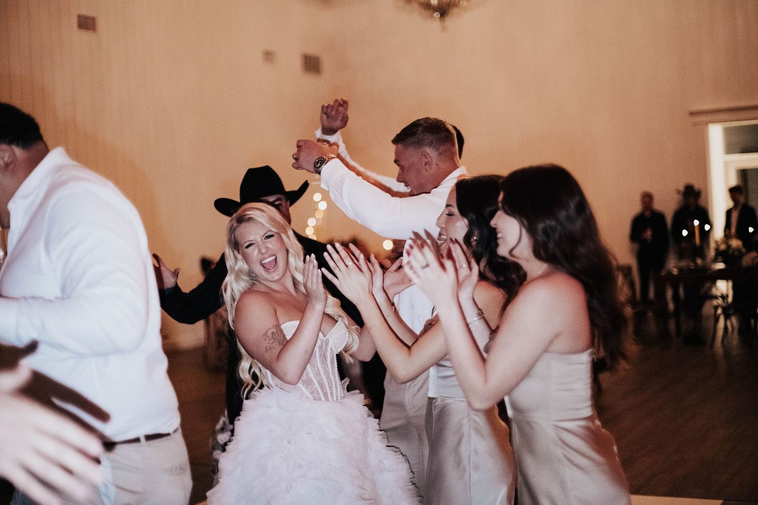 Bride in white dress and cowboy hat celebrates joyfully with bridesmaids and groom at lively wedding reception.