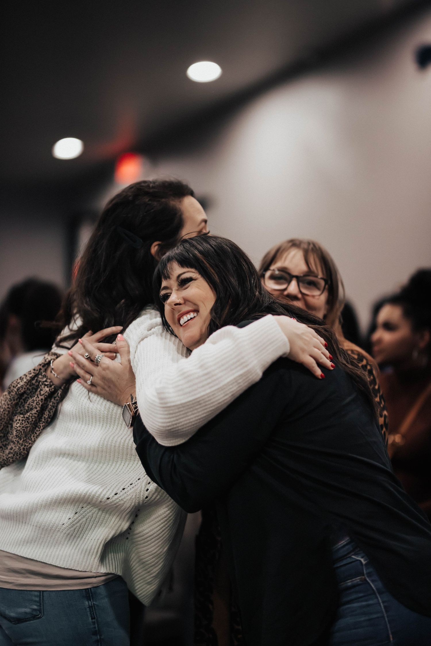 Three women share a warm group hug while smiling at an indoor gathering.