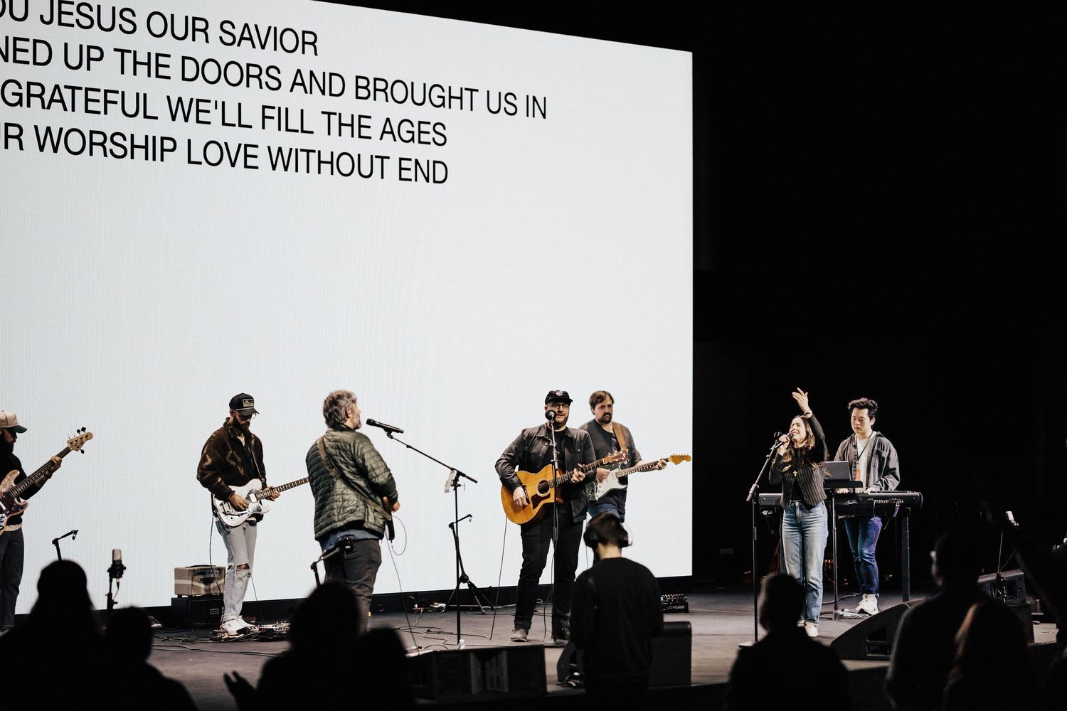 Worship band performs on stage with song lyrics projected on screen behind them.