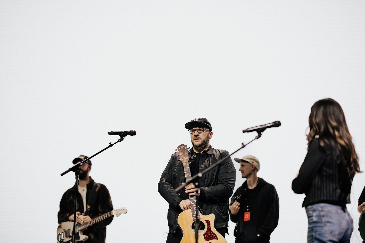 Band performing on stage with guitarist, vocalist holding acoustic guitar, and backup singers against white background.