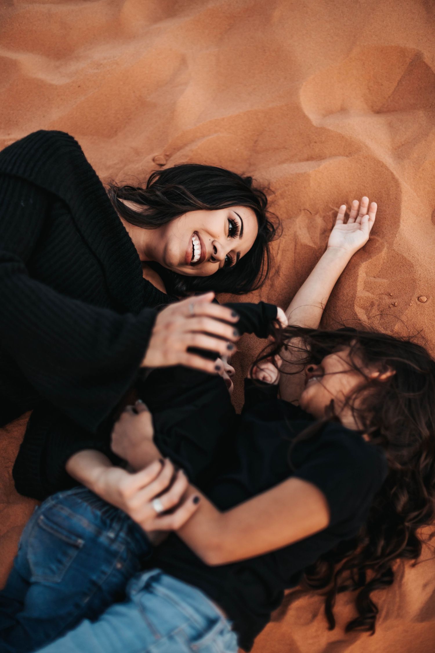 A mother and daughter laughing and playing together while lying on sandy desert dunes.