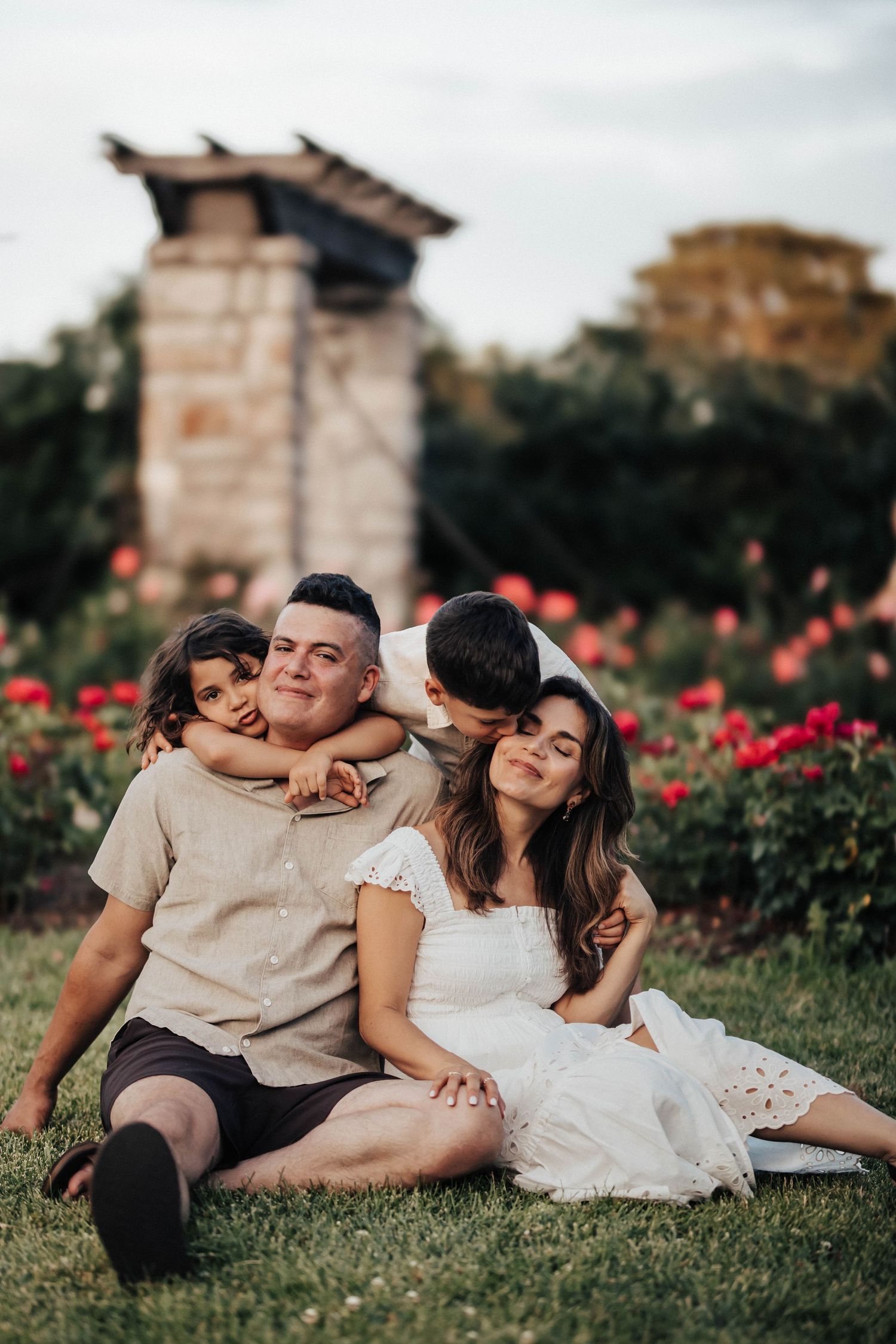 Family of four sitting on grass in a rose garden, parents and two children sharing tender moments outdoors.