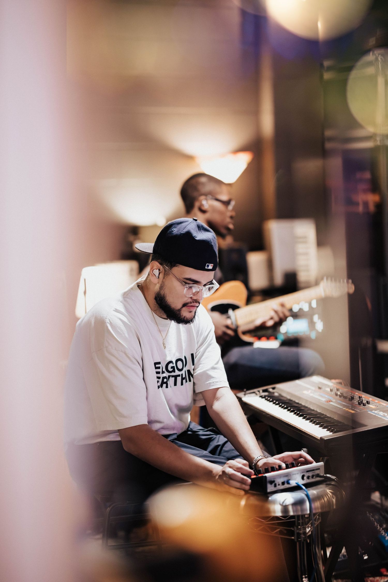 Two musicians in a studio, one playing keyboard with a cap and glasses, another strumming guitar in the background.