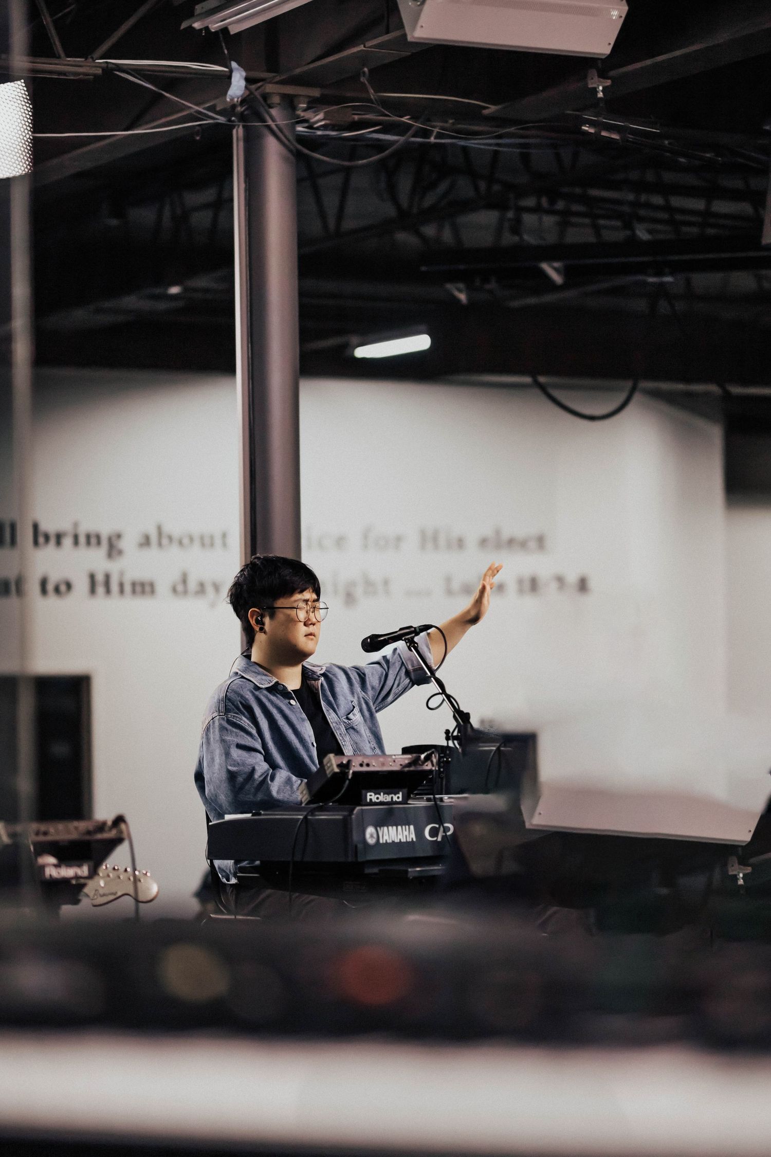 A young Asian musician raises his hand while playing a Yamaha Roland keyboard on stage.