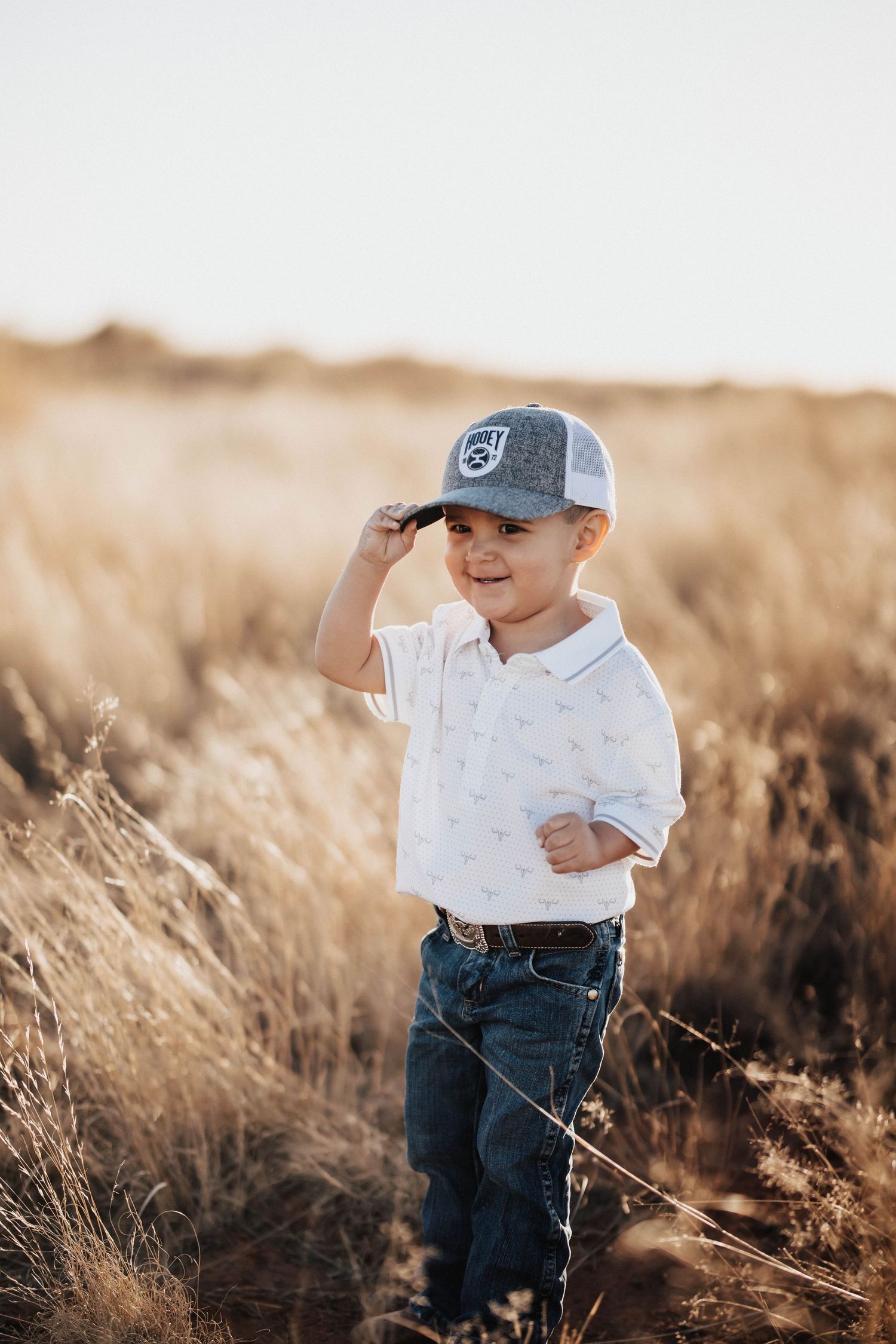 Young boy in white polo, jeans, and gray trucker hat standing in golden field at sunset.