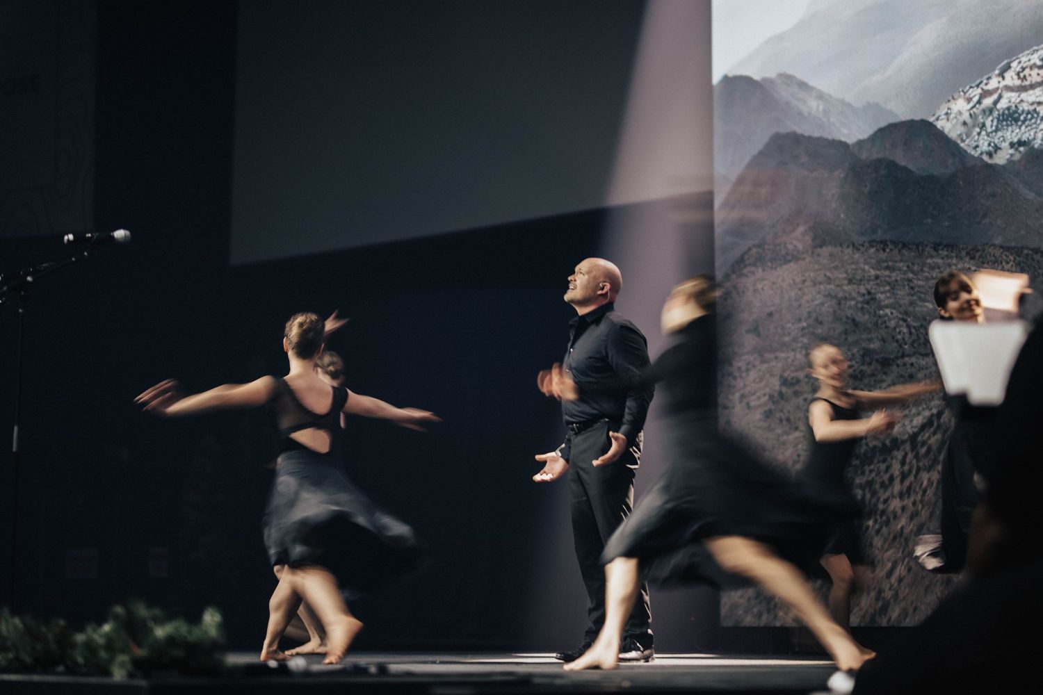 Contemporary dancers perform on stage in front of a mountain projection backdrop under dramatic lighting.