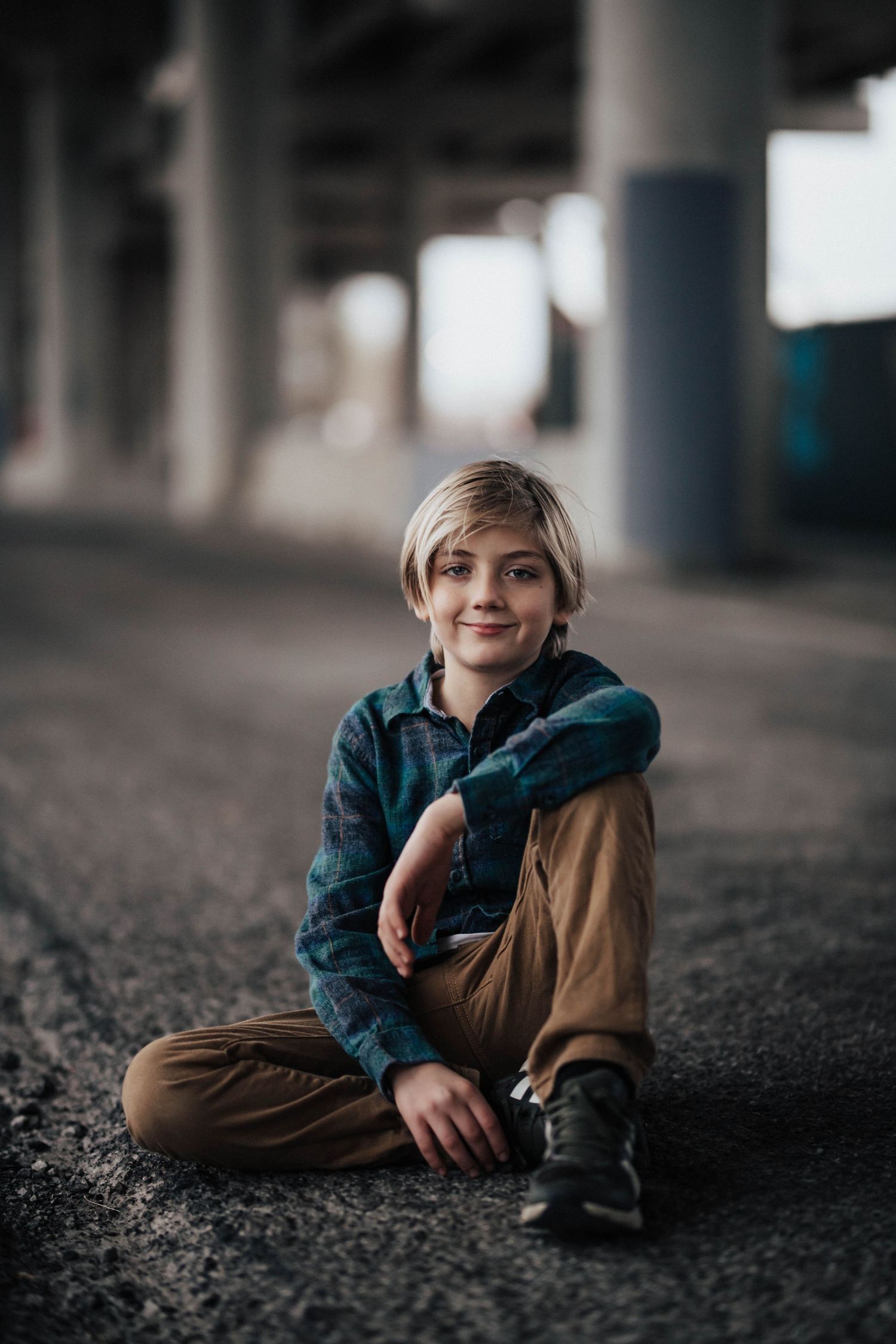 A smiling boy sits cross-legged on pavement under a bridge, wearing a plaid shirt and tan pants.