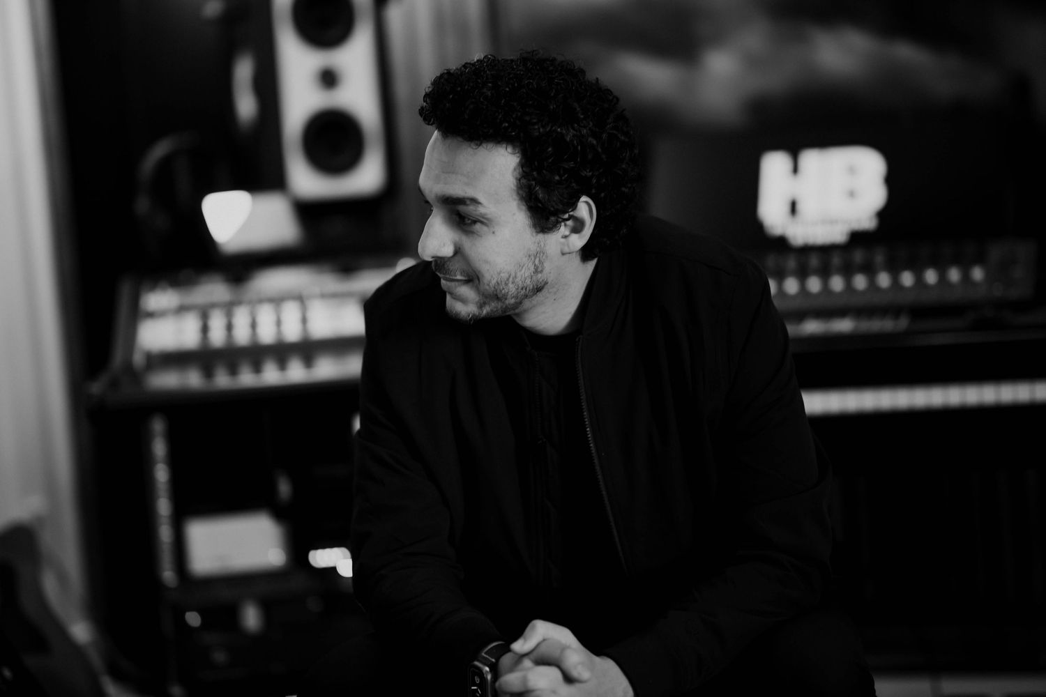 Man in black outfit sits pensively in a professional recording studio with mixing equipment and speakers.