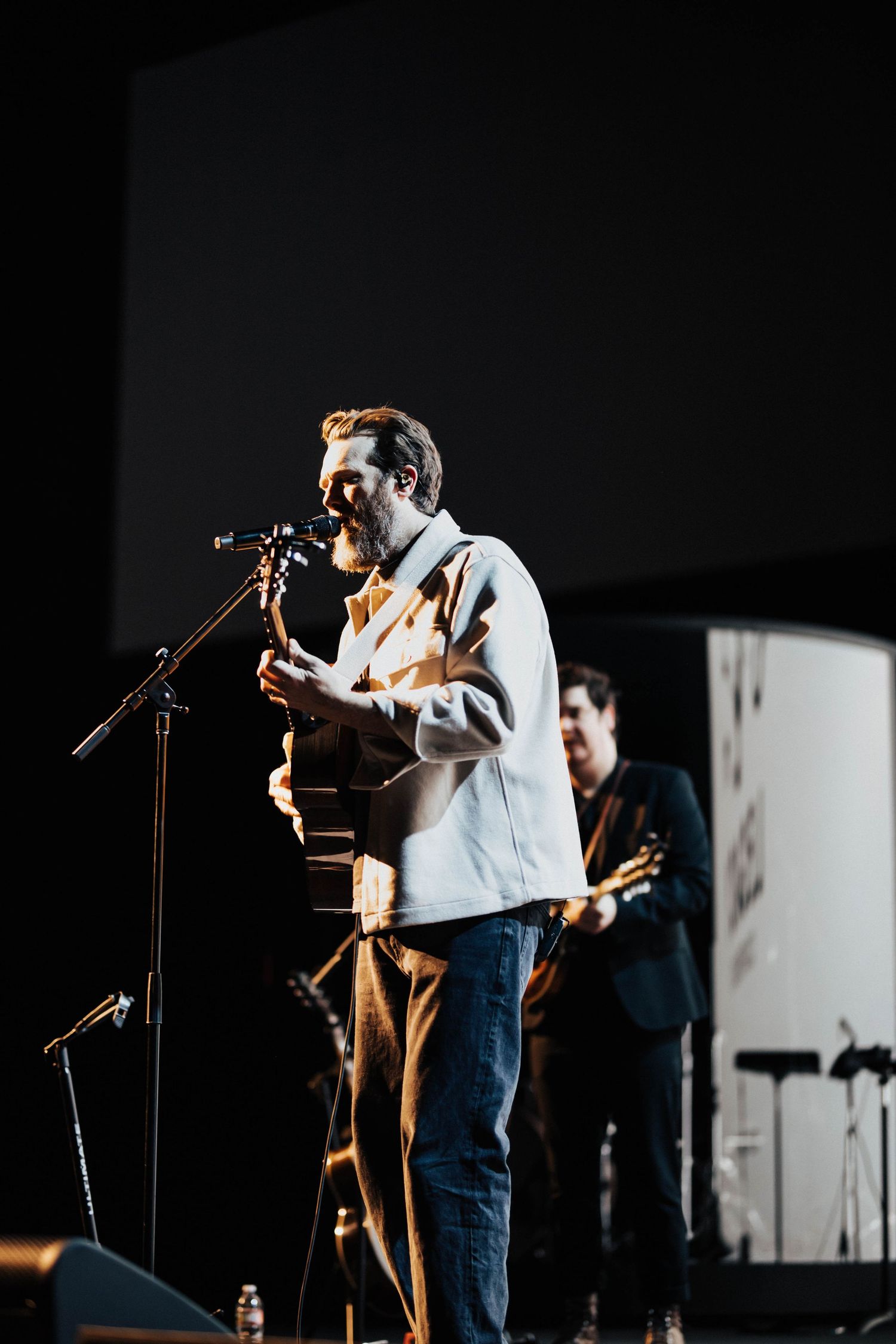 Bearded male vocalist plays acoustic guitar on stage under warm spotlight at live worship concert event.