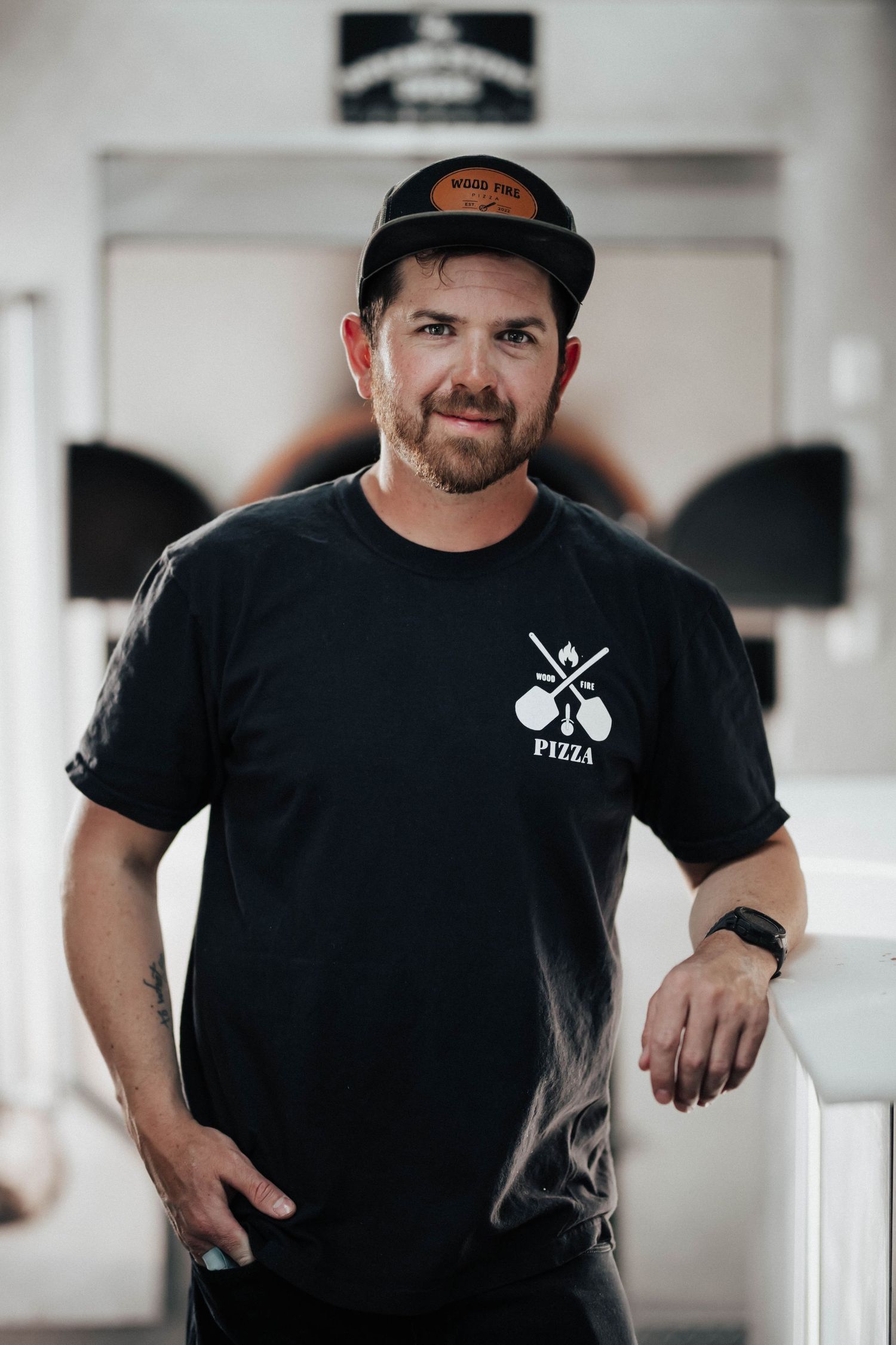 Man in black Wood Fire Pizza t-shirt and cap stands confidently in a wood-fired pizza restaurant.