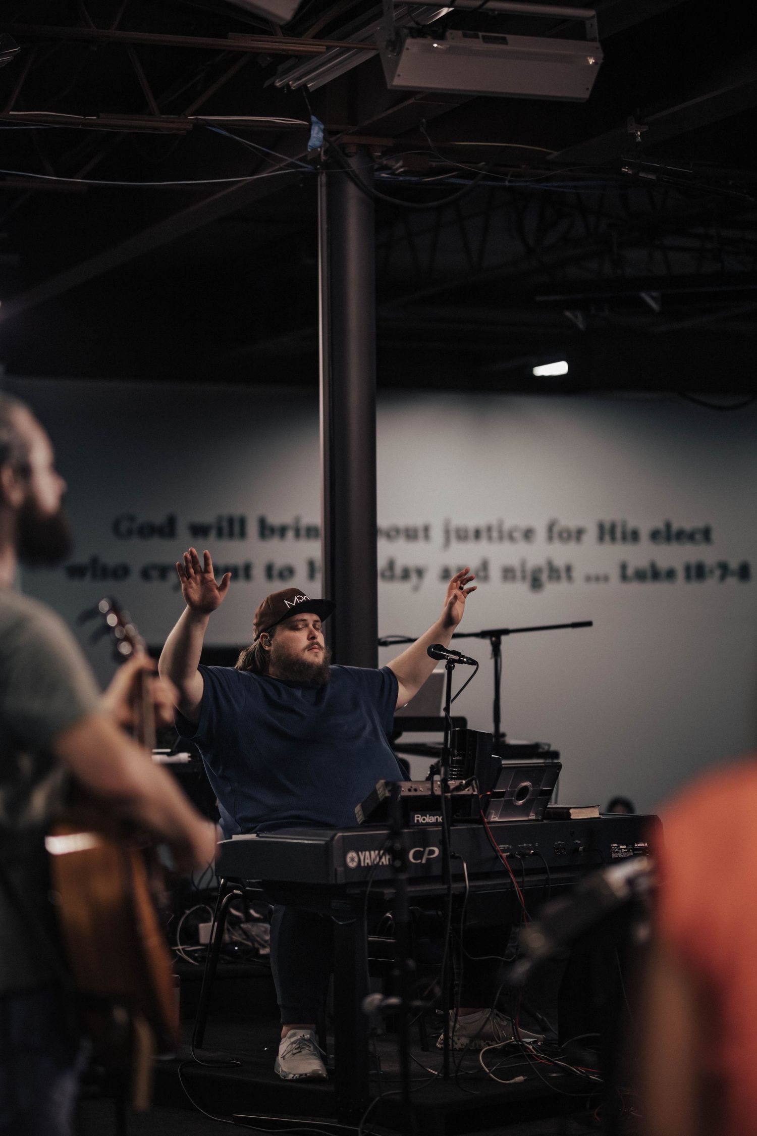 Worship leader raises hands at keyboard during church service with Bible verse on wall.