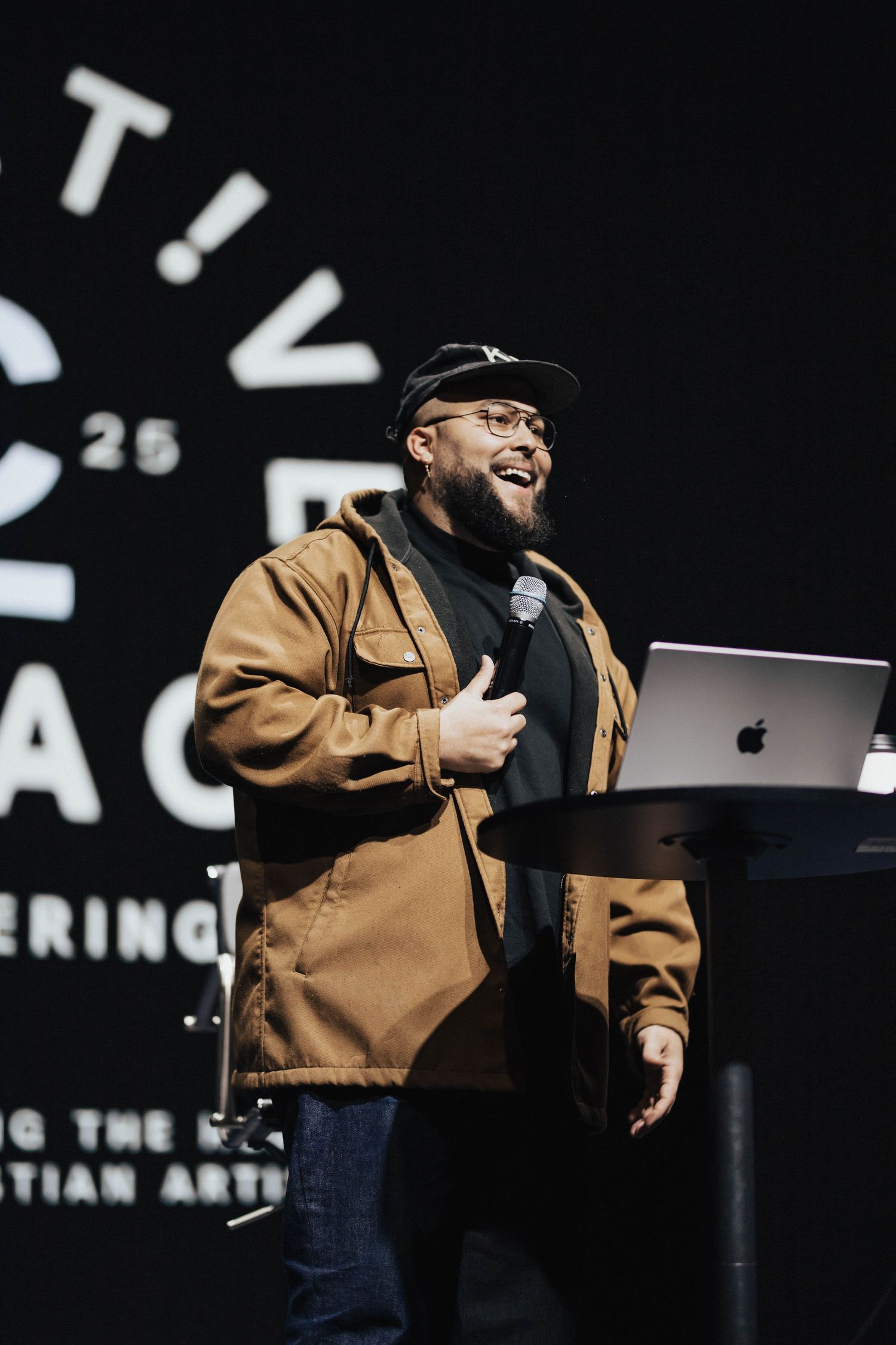 Bearded man in brown jacket and cap speaks on stage holding microphone at conference.