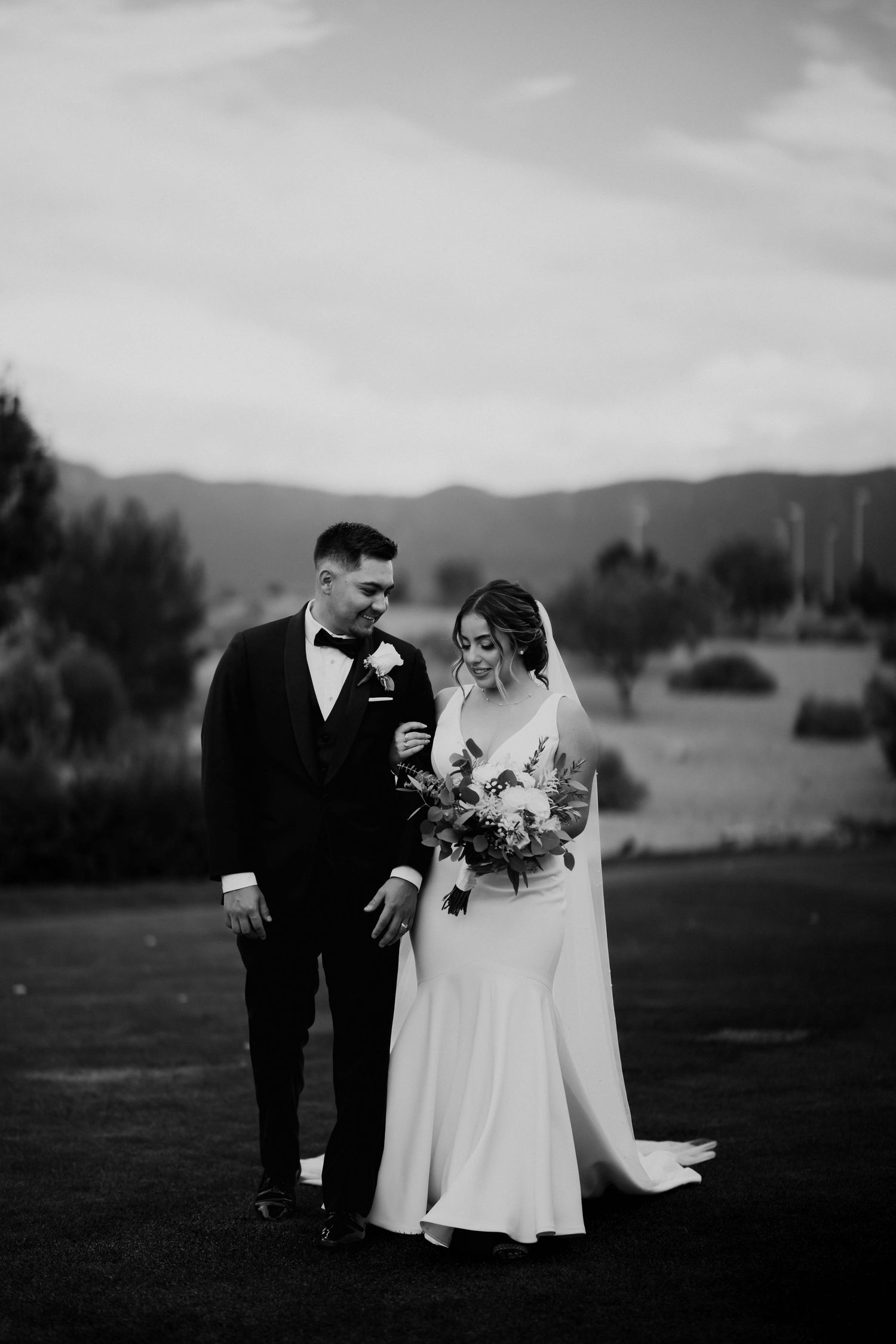 Black and white photo of a bride and groom walking together outdoors with mountains in the background.