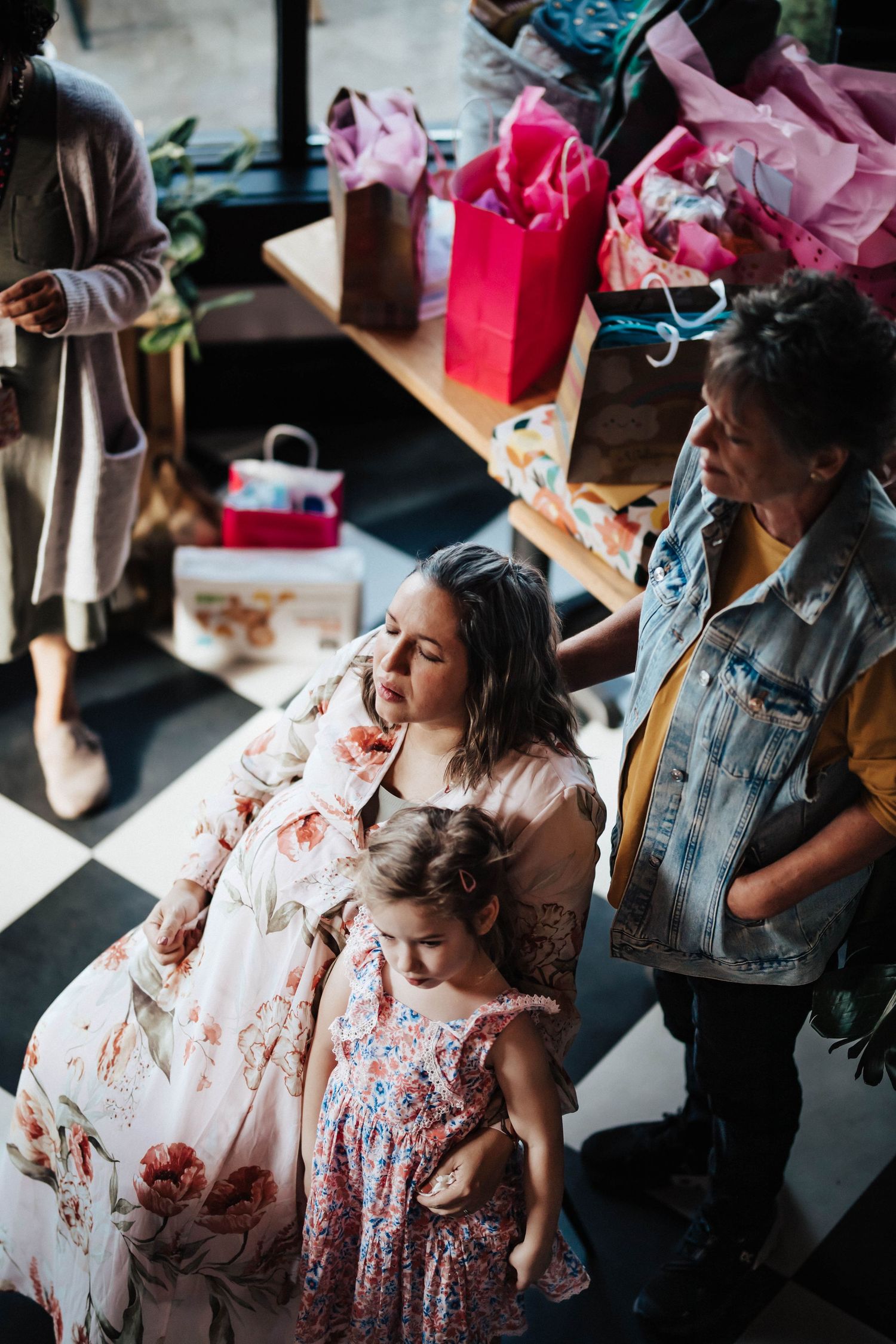 Women and young girl browse colorful gift bags at a baby shower celebration indoors.