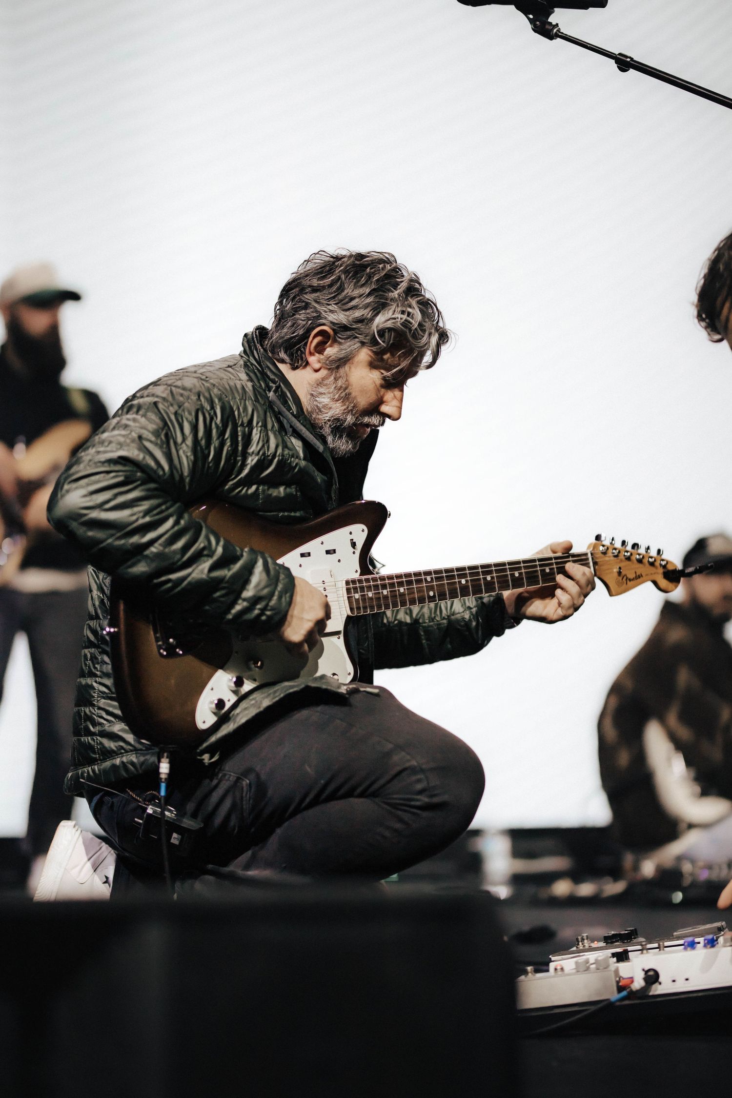 Bearded guitarist in green jacket playing electric guitar while kneeling on stage during live show.
