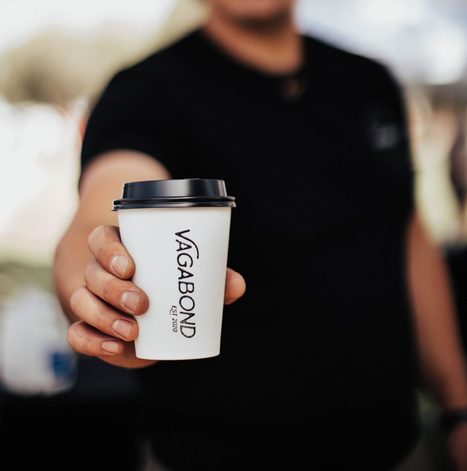 Barista in black shirt extends Vagabond Coffee cup toward camera outdoors.