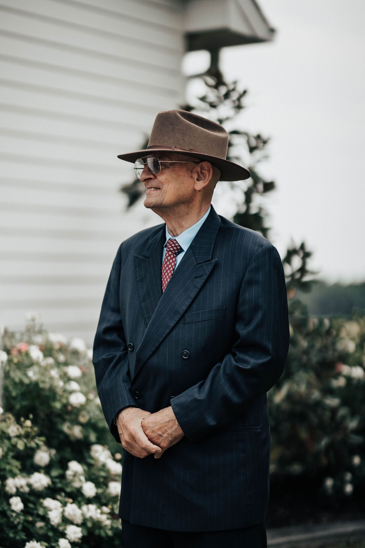 Elegant older man in navy pinstripe suit and brown fedora hat stands outdoors near blooming white flowers.