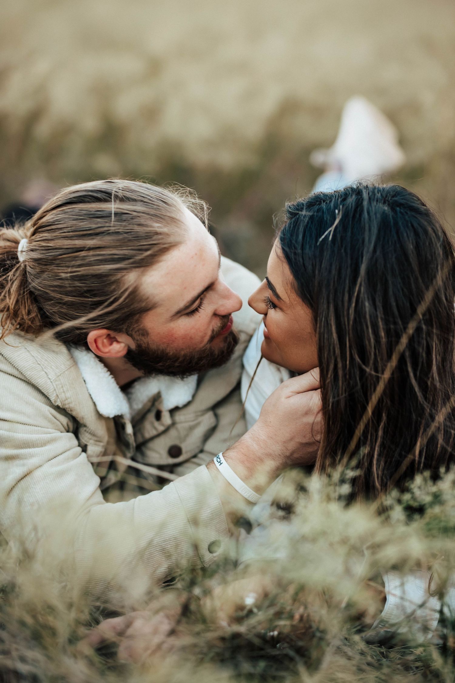 A couple shares a romantic almost-kiss moment while lying together in a field of tall grass outdoors.