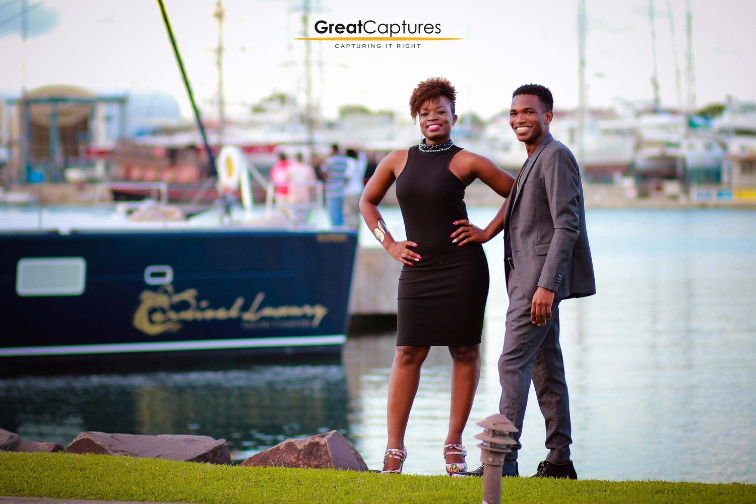 Elegant couple posing by a marina with a luxury yacht, woman in black dress and man in gray suit.