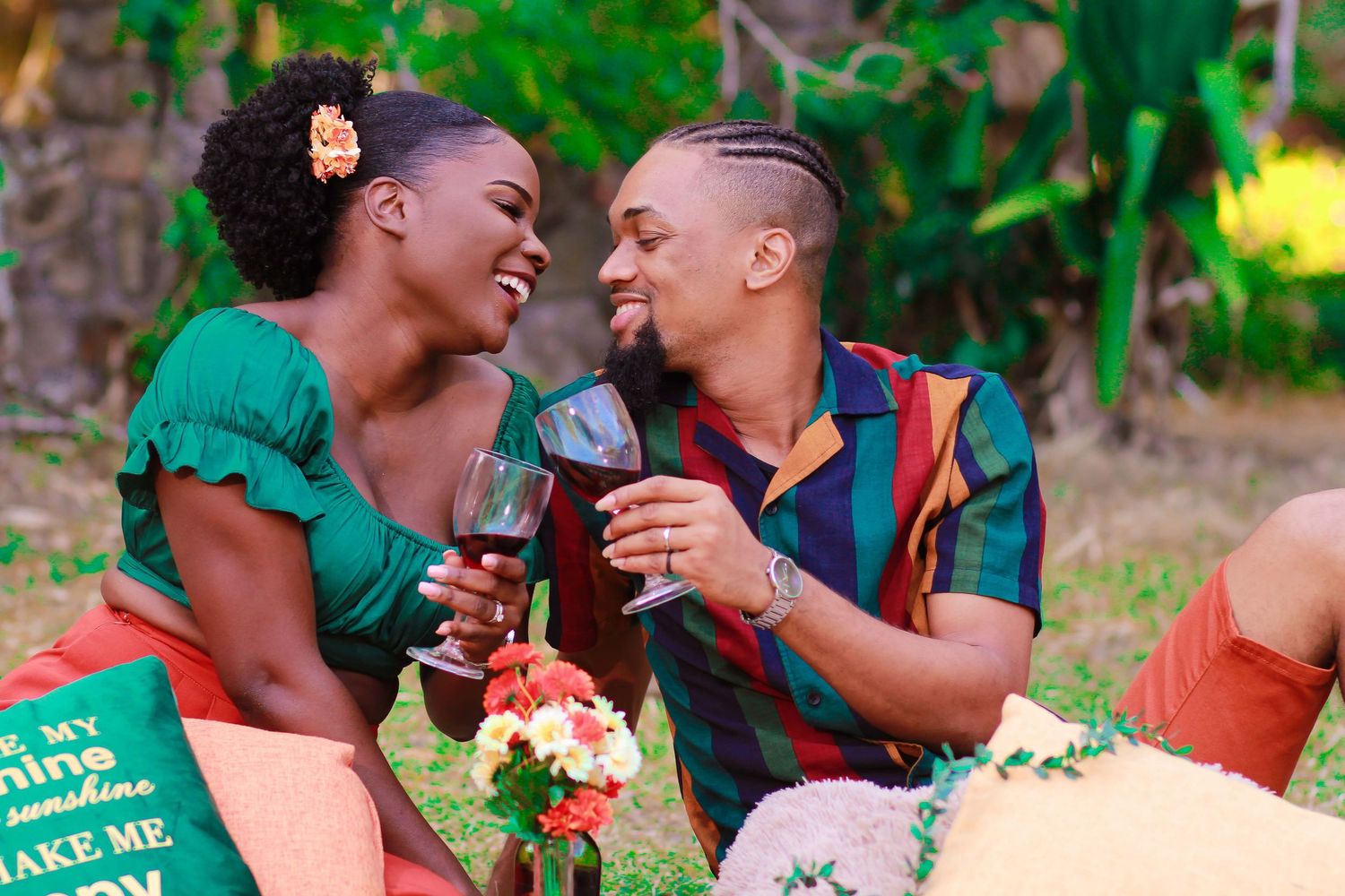 Couple sharing a romantic outdoor picnic, toasting wine glasses and smiling at each other surrounded by flowers.