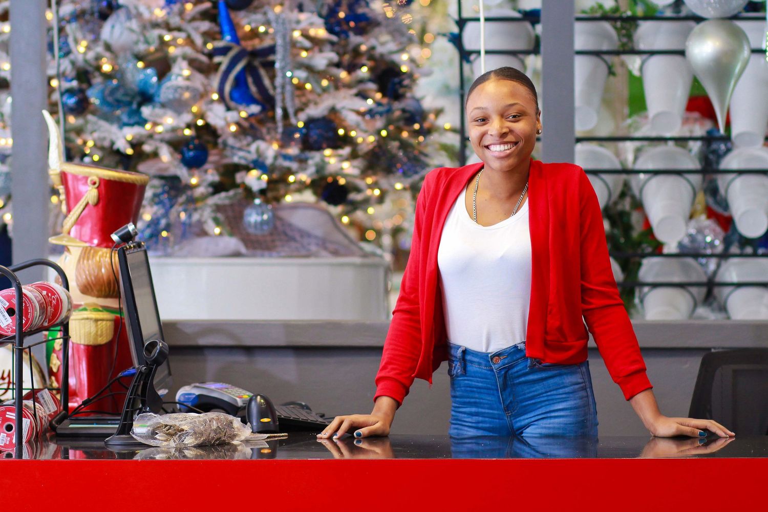 Smiling woman in red cardigan stands at retail counter with festive Christmas tree and decorations in background.