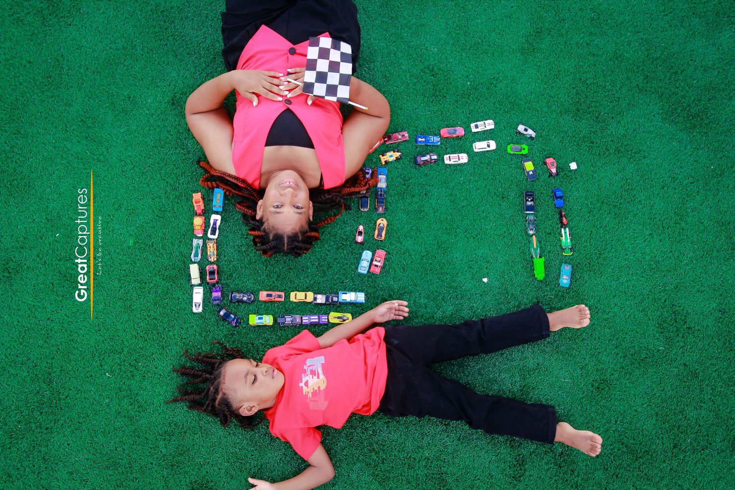 Two children in pink shirts lie on green turf surrounded by toy cars arranged in a creative pattern, viewed from above.