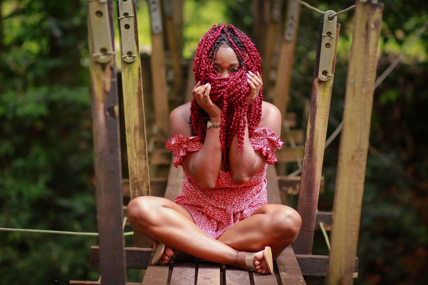 Woman with red braids sitting cross-legged on wooden bridge surrounded by lush green tropical foliage.