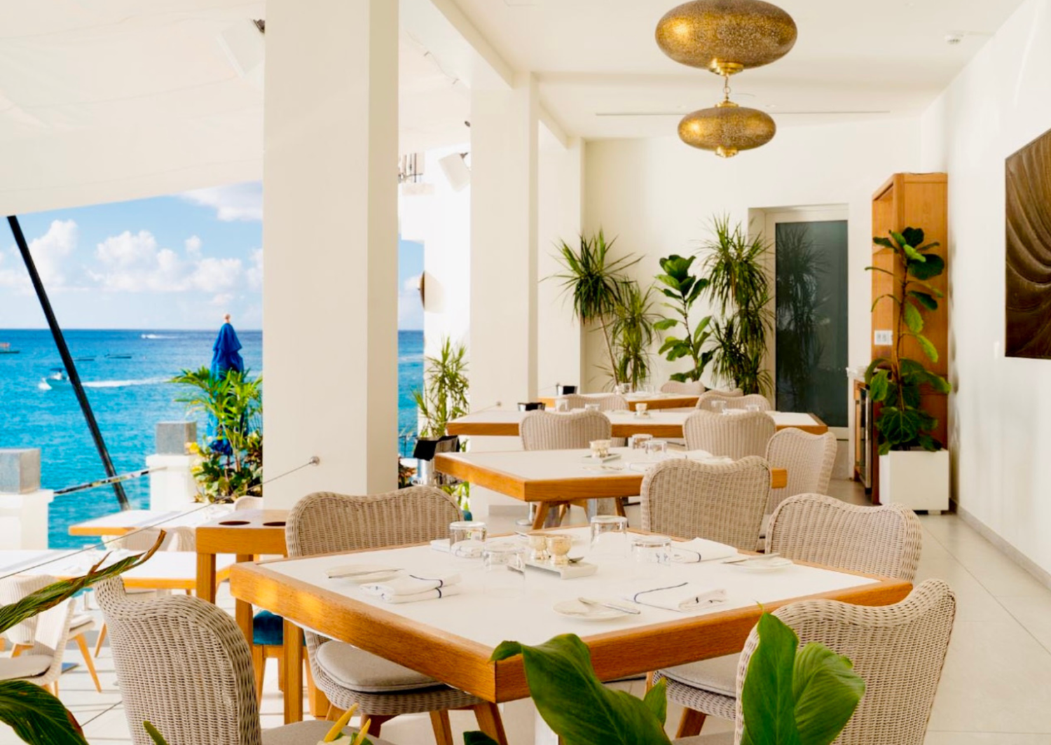 Bright beachfront restaurant with white tables, rattan chairs, gold pendant lights, and stunning turquoise ocean views.